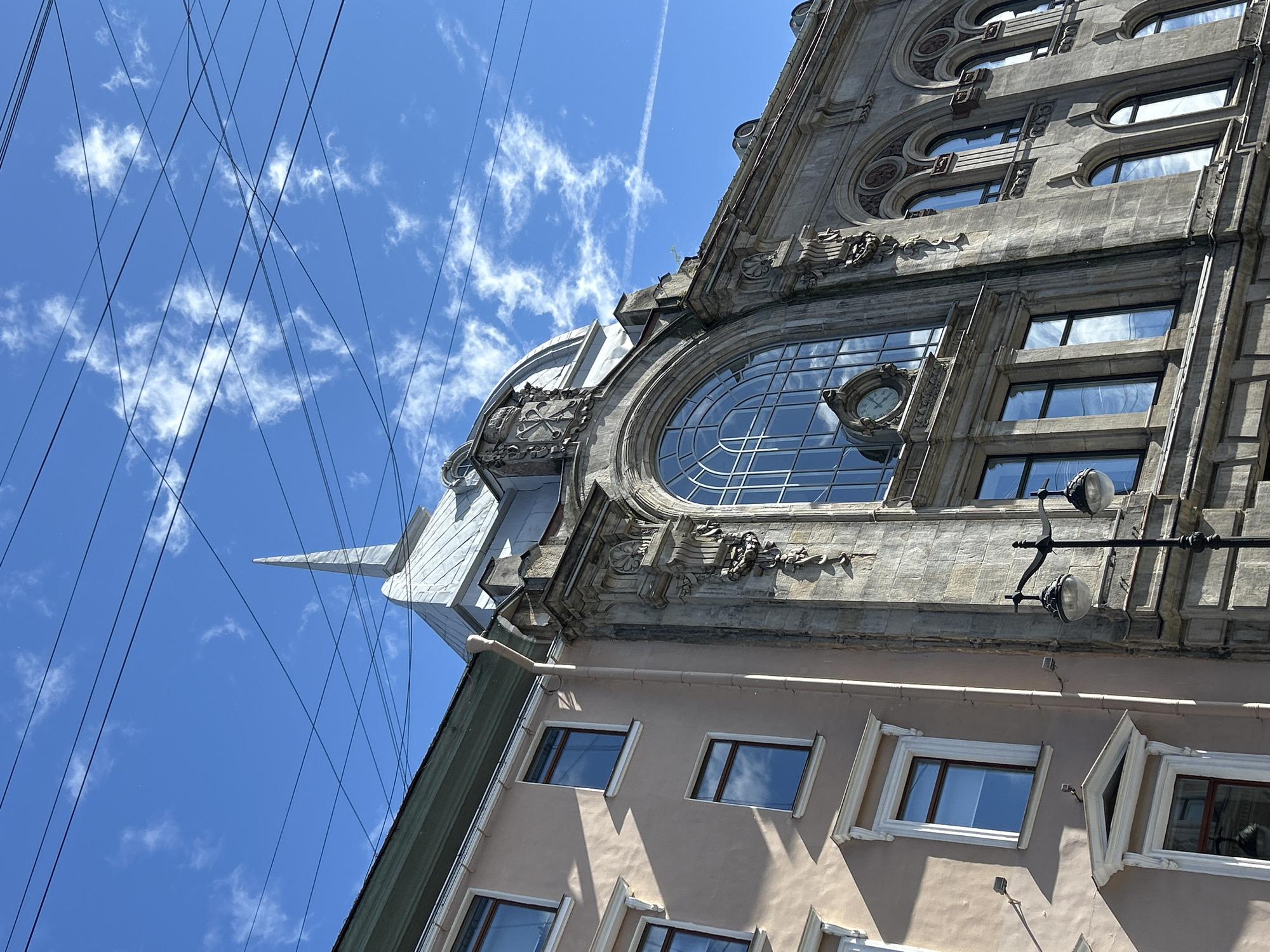 A low-angle, full shot captures the upper portions of two adjacent buildings against a bright blue sky dotted with wispy clouds and crossed by numerous overhead wires. The building on the left, painted a pale peach color, features several regularly spaced windows with dark frames. Decorative white moldings accentuate the tops of some of these windows. The building on the right is constructed of stone with ornate architectural details. Its most prominent feature is a large, arched window with a grid of panes, and within this window, a clock face is visible. Below this, a row of smaller rectangular windows with dark frames are present. A steeply sloped, silver-colored roof with a pointed spire is visible between the two buildings, extending upwards. A black lamppost with two globe-shaped lights stands in the foreground, partially obscuring the lower part of the stone building. The lighting suggests it is daytime.