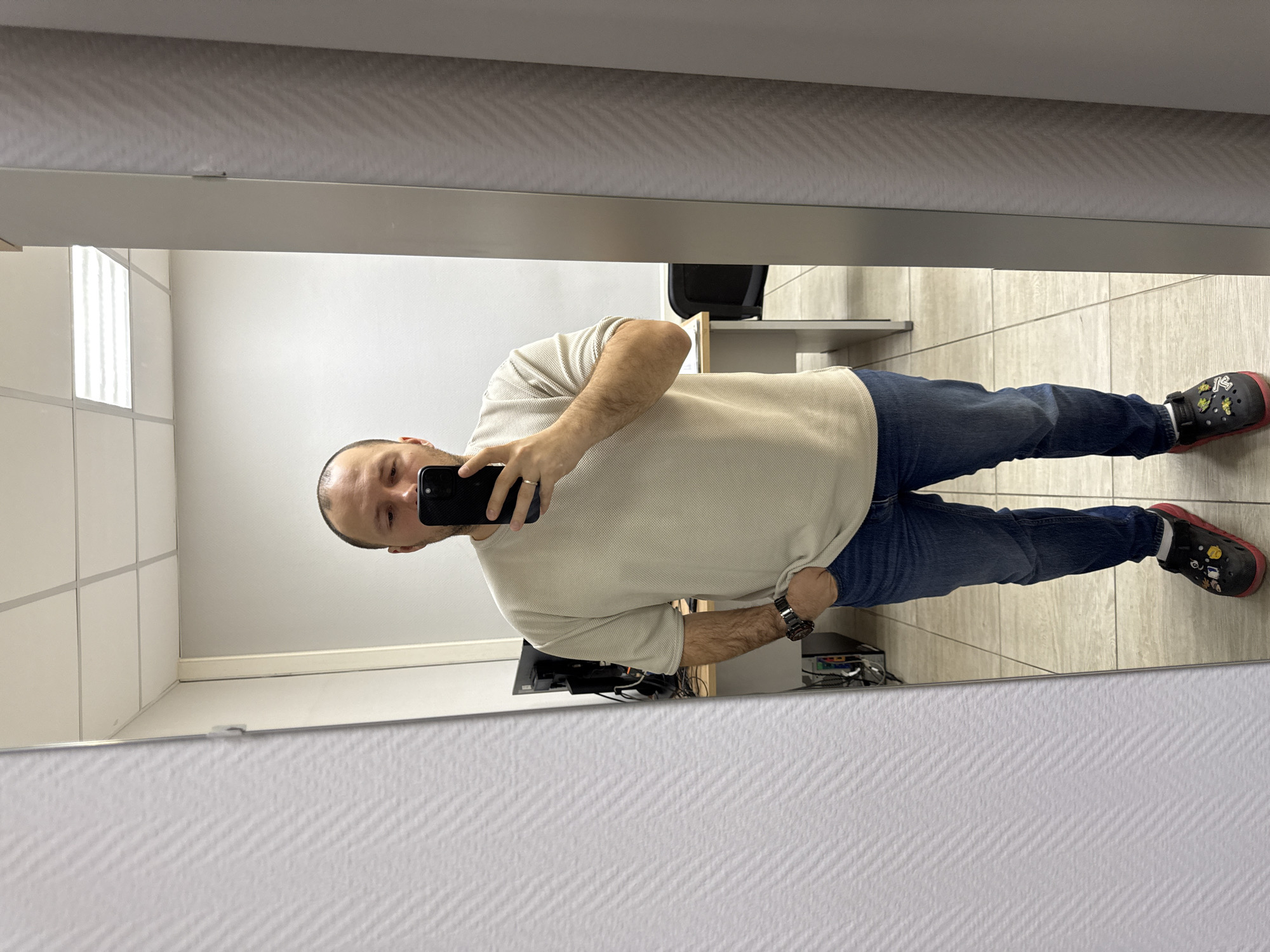 A man is taking a selfie in a mirror. He is wearing a light-colored short-sleeved shirt, blue jeans, and Crocs with red soles. He is holding a black smartphone in his right hand. The reflection shows a room with a tiled floor and a drop ceiling. A desk with computer equipment is visible behind him. A chair is positioned against the wall. The exact location in Krasnoyarsk, Russia, cannot be determined from the image alone.