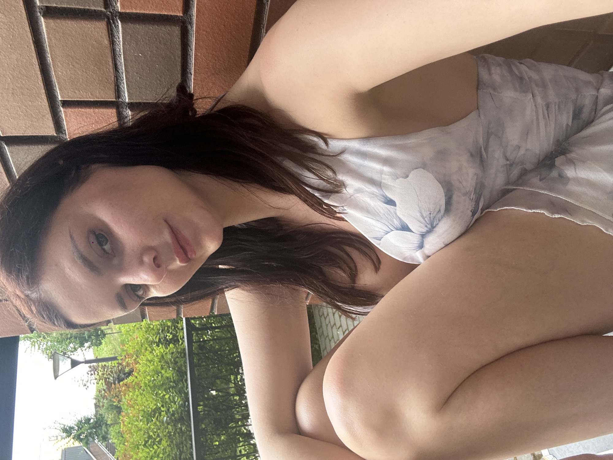A young woman is captured in a close-up, sideways shot, her head resting on her arm. She has long, dark hair and is wearing a light-colored garment with a floral pattern. Her eyes are open, and she appears to be looking directly at the camera, with a slight smile on her lips. The background is out of focus, but hints of an outdoor setting are visible, including greenery and what looks like fencing. The lighting suggests it might be daytime. The image is somewhat tilted, adding a casual, candid feel to the pose.