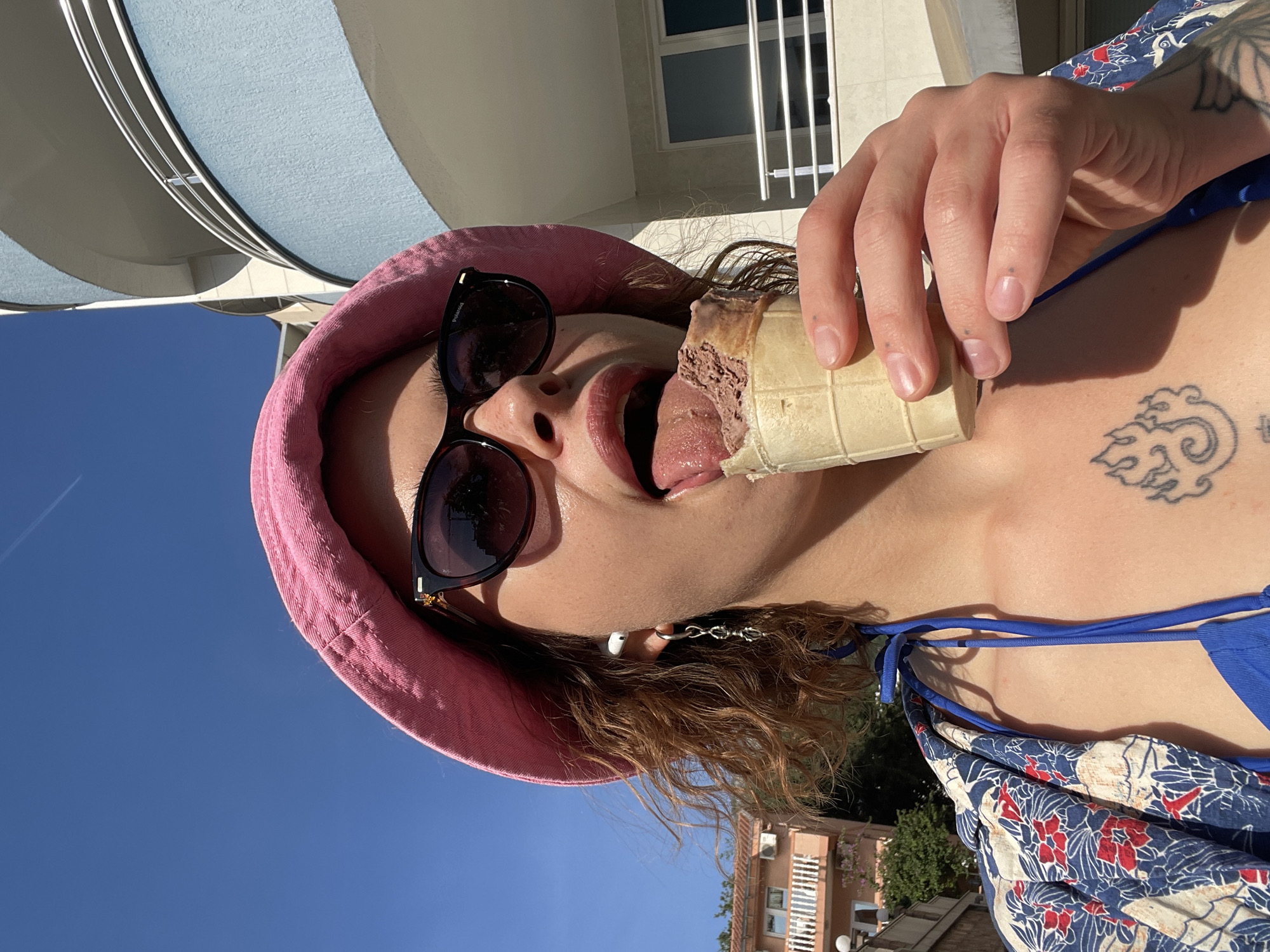 The image shows a person in Rafailovići, Montenegro, wearing a pink hat and sunglasses, holding an ice cream cone and licking it with their tongue. They also have a tattoo on their chest. The background includes buildings and the sky.