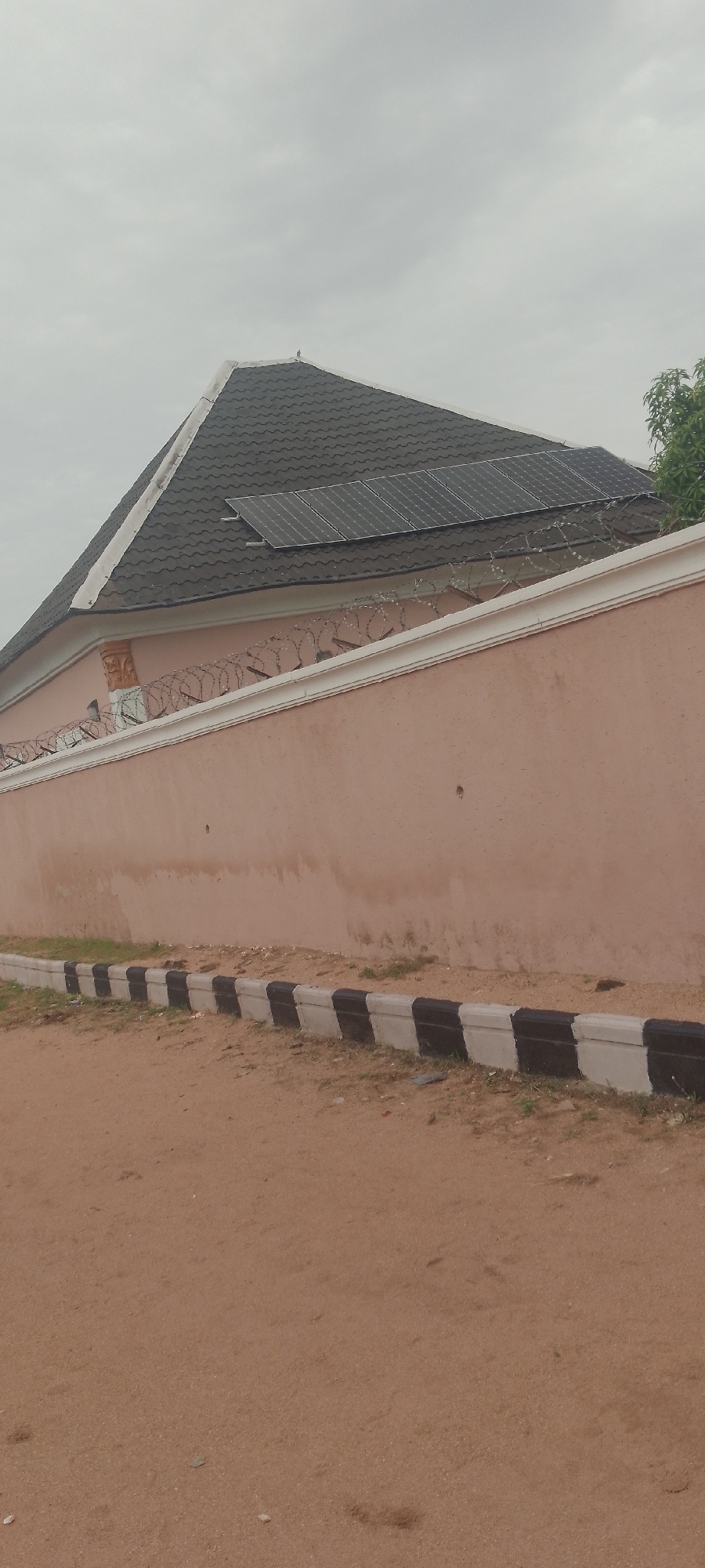 A drone shot from an elevated position captures a residential building in Jalingo, Nigeria, on an overcast day. The building features a dark, shingled roof with several solar panels installed on one side. A pale pink wall with barbed wire running along its top delineates the property. The wall is bordered at the bottom by a strip of black and white painted concrete blocks, suggesting a decorative or protective edge. The ground in front of the wall is a reddish-brown dirt with sparse patches of dry grass. To the right of the frame, a lush green tree stands out against the muted sky. A tiny bird is perched on the peak of the roof, barely visible against the grey expanse. The scene appears quiet and undisturbed, with no visible human presence or activity.