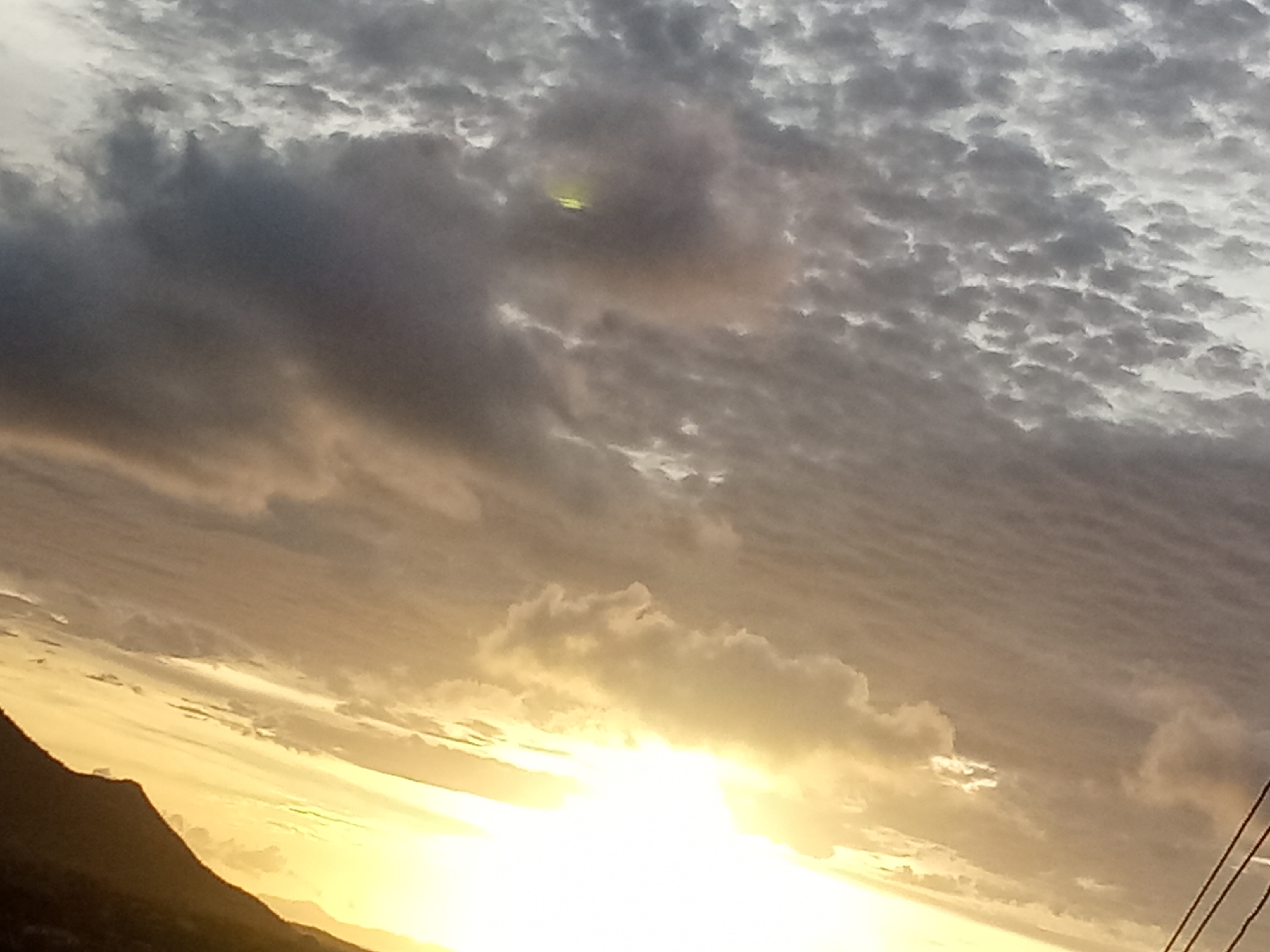 The image depicts a sunset in what appears to be a mountainous area, possibly near Altagracia, Venezuela. The sky is filled with a mix of dark gray and light, altocumulus clouds. The sun is positioned low on the horizon, emitting a bright, golden light that illuminates the clouds and landscape. A dark silhouette of a mountain is visible in the lower left corner of the image. There are no visible people, actions, or interactions present in the scene. The environment is dominated by the sky, clouds, sunlight, and mountain silhouette. The focus of the image is on the sunset and the atmospheric conditions.
