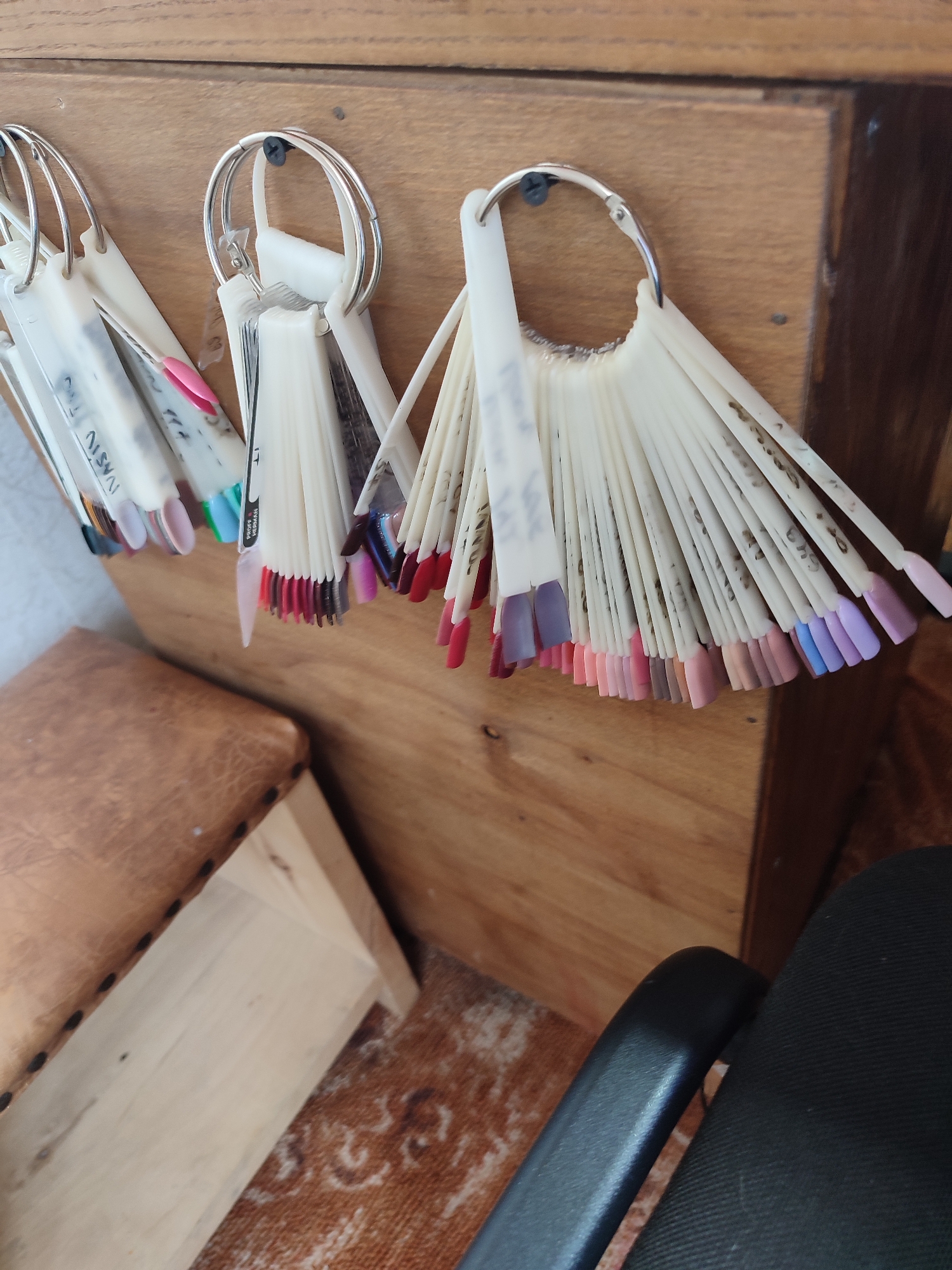 An indoor, eye-level, medium shot captures a wooden shelf displaying fan-shaped nail polish swatches. Three metal rings, each holding multiple swatches, are attached to the shelf with screws. The swatches vary in color, ranging from light pinks and purples to darker reds and blues. Some swatches have handwritten labels on them. To the left of the shelf, a portion of a light brown, leather-covered stool with black decorative tacks is visible. The floor beneath the stool is covered with a patterned rug. To the right of the shelf, the armrest and part of the seat of a black office chair are visible. The background consists of a light brown wooden surface. The location is Yaresky, Ukraine.
