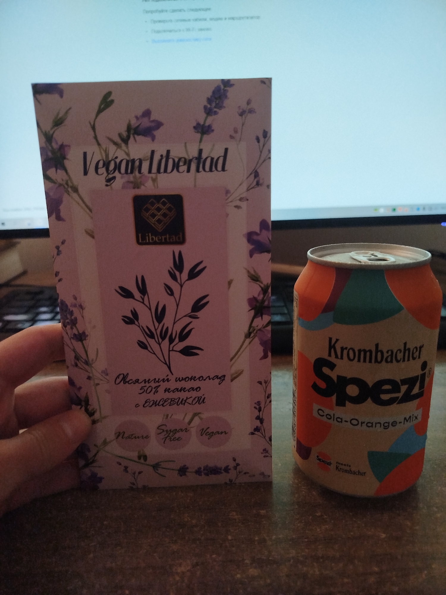 An overhead shot captures two items on a speckled brown surface, likely a desk. To the left, a rectangular box of "Libertad" vegan chocolate is held at an angle by a person's hand, which is visible from the knuckles down. The chocolate box features a light purple background with a floral pattern and the text "Vegan Libertad" at the top. Below the logo, a sprig illustration is prominent, and text in Cyrillic script indicates "Oвсяный шоколад 50% какао с ежевикой" (Oat chocolate 50% cocoa with blackberry), along with icons for "Nature," "Sugar Free," and "Vegan." To the right, a Krombacher Spezi cola-orange mix soda can stands upright. The can's design includes colorful abstract shapes and text. In the background, a computer monitor displays a blurred image of a webpage with text, suggesting an indoor setting.