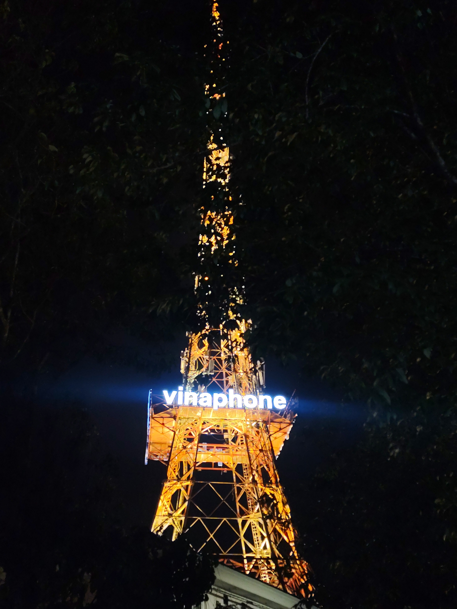 The image shows a night-time view of a tall radio tower in Da Lat, Vietnam. The tower is illuminated with yellow and orange lights, creating a vibrant contrast against the dark night sky and surrounding trees. At the top of the tower, a bright blue neon sign reads "Vinhaphone." The tower structure is intricate, made of crisscrossing metal beams. The overall scene is dimly lit, with only the tower and its signage being brightly lit.