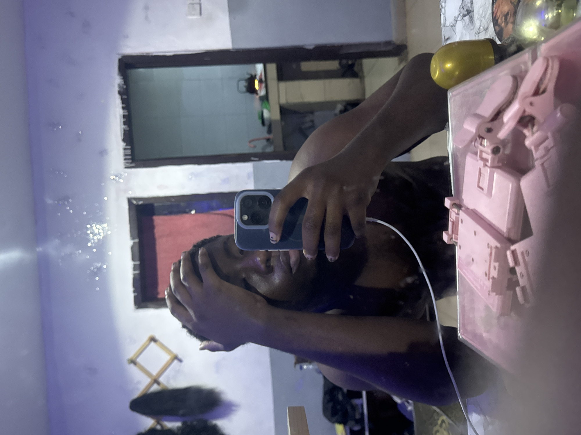 A man is taking a selfie in front of a mirror. The reflection in the mirror shows a room with a doorway leading to another room. A folding wooden stand and dark-colored items are visible in the lower left of the reflection. A pink object is prominent in the foreground, to the right of the man's reflection. The location is Ipetumodu, Nigeria.