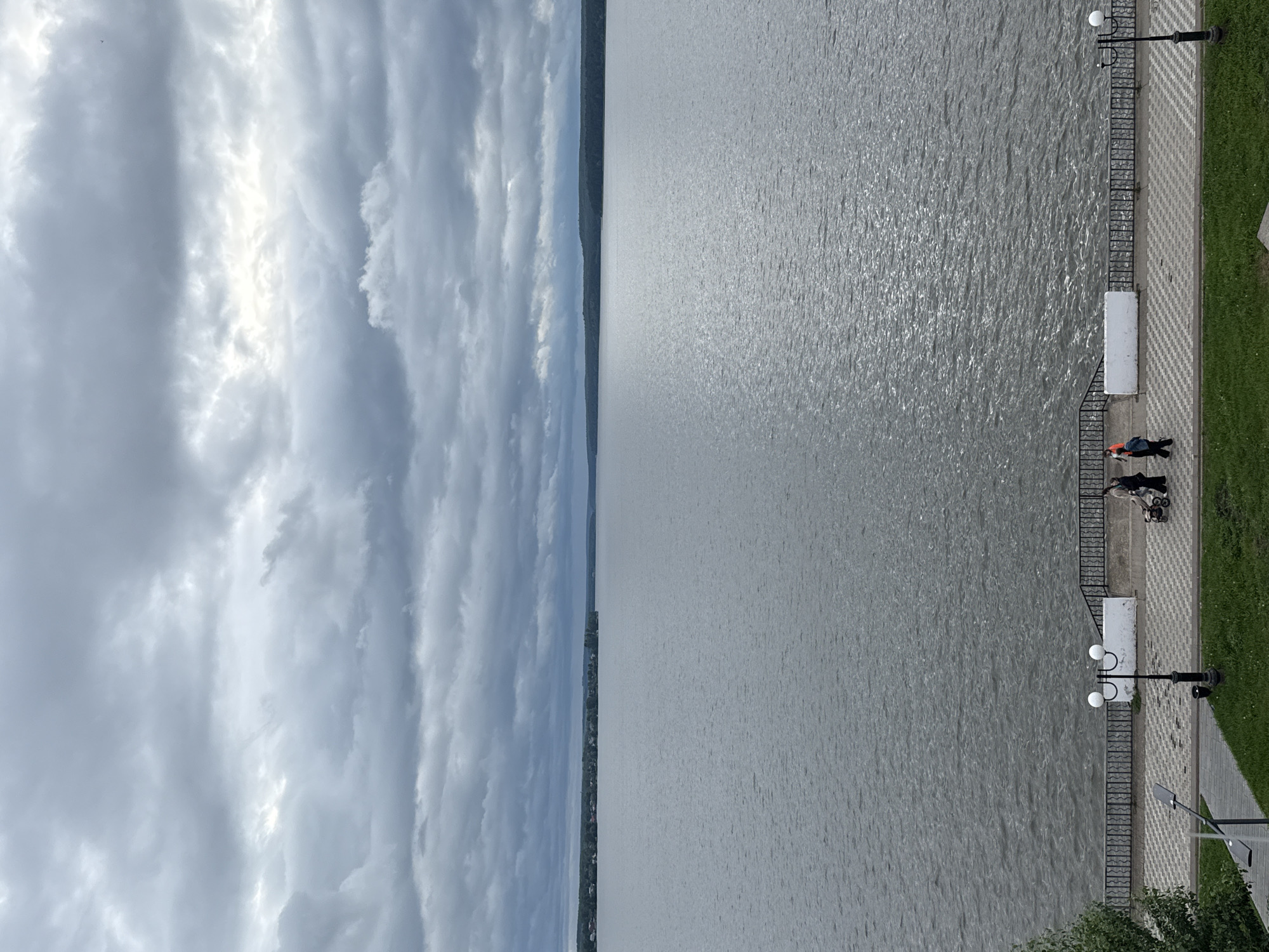 This image presents a broad view of the Votkinsk Reservoir in Votkinsk, Russia, under an overcast sky. The scene is bisected by the horizon line, separating a cloudy sky from a large body of water.
The upper portion of the image is filled with a dense layer of grey clouds, displaying variations in tone from dark grey to lighter patches where some illumination penetrates.
The central part of the image features the wide expanse of the Votkinsk Reservoir, its surface appearing a uniform grey with subtle ripples and faint reflections. The distant shore is visible as a low-lying landmass covered with trees. On the left side of the far shore, scattered structures suggest a small settlement, while the right side of the distant shore is characterized by an unbroken line of dense forest.
In the foreground, a paved promenade runs along the water's edge. The promenade features a checkered pattern of light and dark grey tiles. A black metal railing separates the walkway from the water. Two white rectangular structures, possibly benches or decorative elements, are situated behind the railing. Two black lampposts with white spherical lights are positioned on the promenade, one in the mid-left and another on the far right. A modern street light fixture is partially visible on the far left. Below the promenade, a manicured lawn of green grass slopes upwards from the bottom of the frame, with a portion of a leafy green tree visible on the extreme left.
Three individuals are present on the promenade. On the right side, two people are walking towards the left. One individual, dressed in dark clothing, pushes a baby stroller. The second individual, walking alongside, wears dark clothing with a bright orange upper garment or accessory. A third person, clad in dark attire, is partially visible on the left side of the promenade near the mid-left lamppost, facing away from the camera.