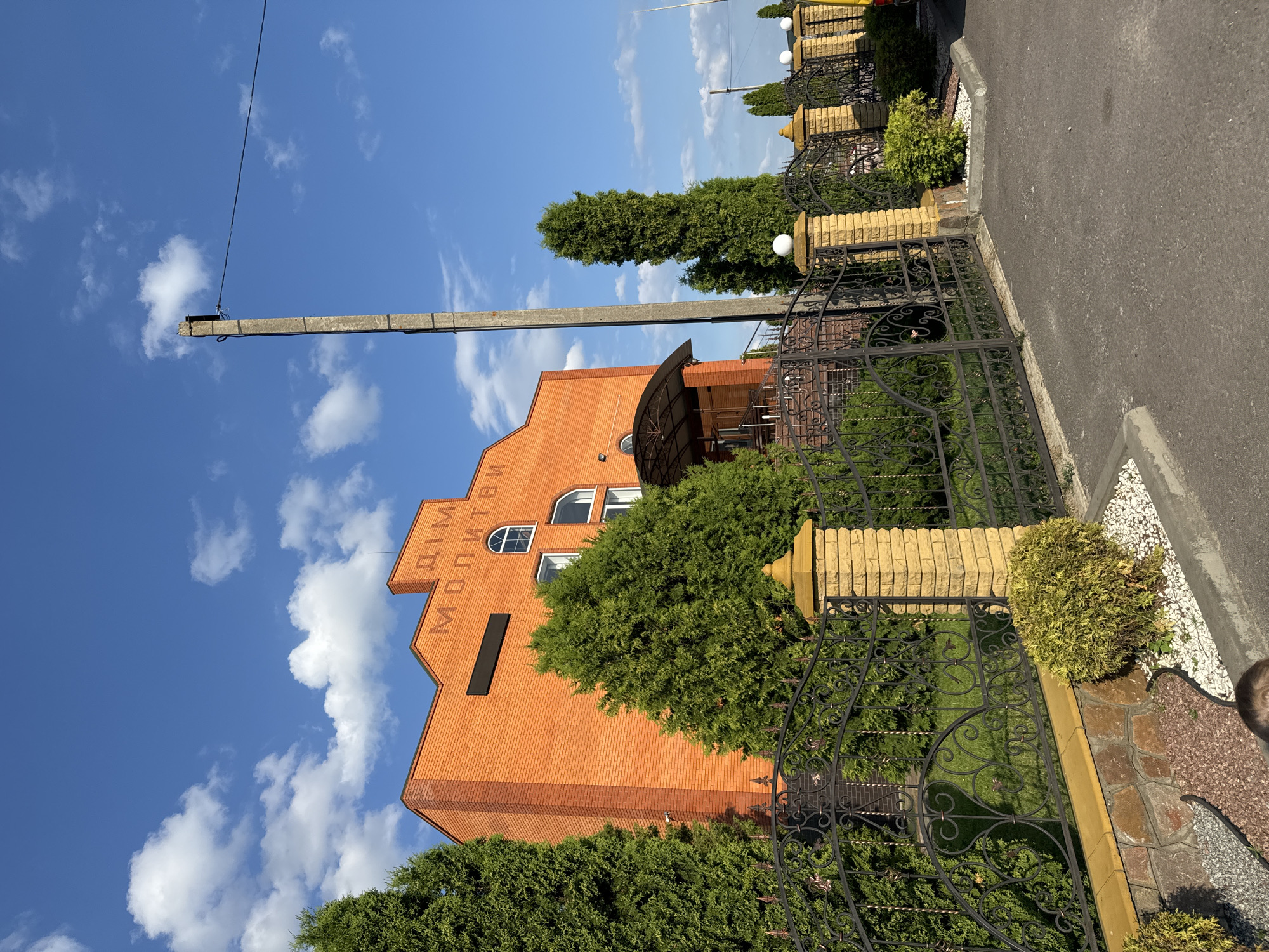 The image shows a bright orange brick building with the words "ДІМ МОЛИТВИ" (House of Prayer) inscribed on its facade. The building is situated behind a decorative black metal fence with yellow brick pillars topped with finials. Evergreen trees flank the building and the fence. A concrete utility pole stands to the right of the building. The sky is blue with scattered white clouds. The scene appears to be a street in Dovhe, Ukraine.