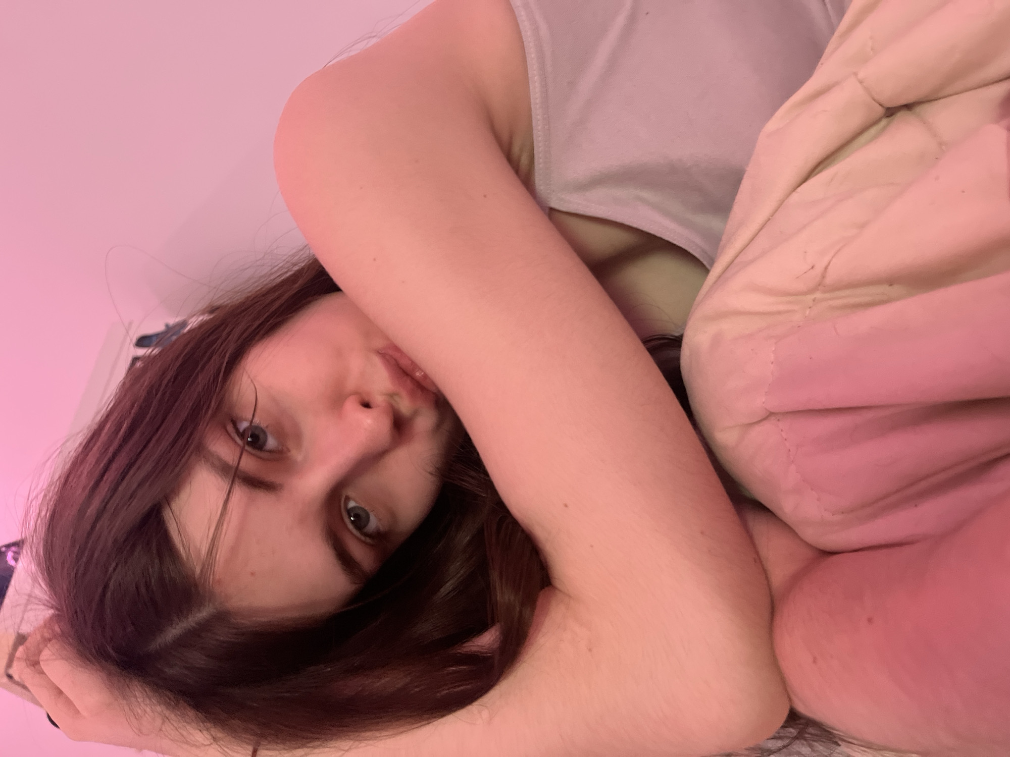 A young woman with long brown hair and green eyes lies in bed with a light-colored blanket pulled up around her. She is wearing a light-colored tank top. The lighting in the room has a pink hue. There is no indication of the location being Saint Petersburg, Russia.