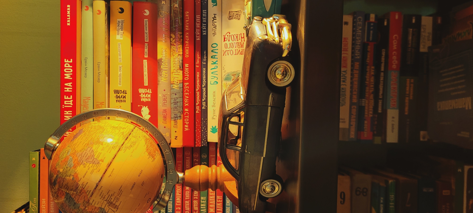 A tabletop scene features a vintage globe and a model car placed on a bookshelf filled with books. The globe is illuminated, casting a warm glow. The model car, a dark-colored coupe, is positioned in front of the books. The bookshelf itself is packed with books of various sizes and colors, with visible titles in Cyrillic script. The background is a plain green wall. The scene appears to be indoors, possibly in a home or study.