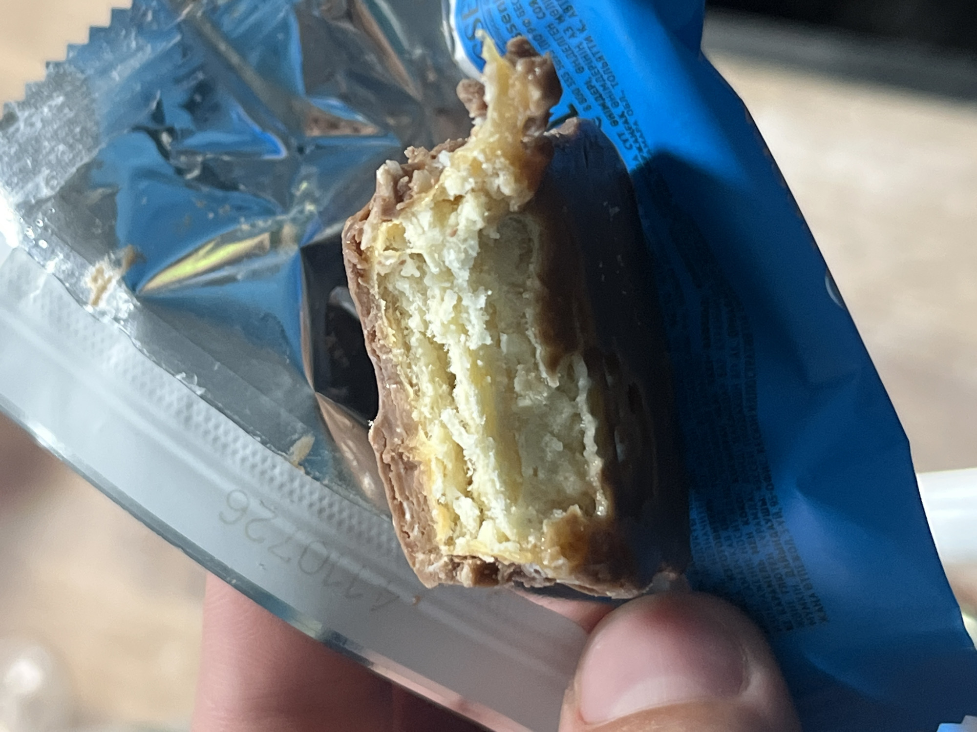A hand holds a partially eaten chocolate bar, its nougat-like interior exposed. The bar is wrapped in crinkled silver foil and a blue plastic wrapper. The blue wrapper has text printed on it in a foreign language, likely Russian, given the location context. Some of the text is readable, including what appears to be an expiration date or batch number: "110726". The scene is a close-up shot, with the background blurred. The lighting suggests it is daytime, likely indoors. No specific emotions are evident.