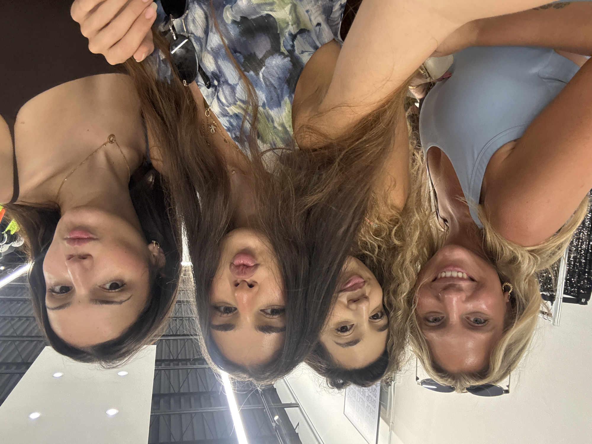 Four women are closely positioned in a selfie taken from a low angle. The woman on the far left has dark hair and wears a brown top with a delicate gold necklace. Next to her is a woman with long, straight, brown hair, and floral dress. Another woman is beside her, also with long brown hair, styled with curls. The woman on the right has blonde hair, wears a light blue top, and black sunglasses. The women are indoors with a ceiling featuring fluorescent lights visible in the background, and clothes racks. Based on the "SHOP NEXT" sign appearing to the left, the subjects are inside of a shop. There is no way to identify the exact location of Miami.