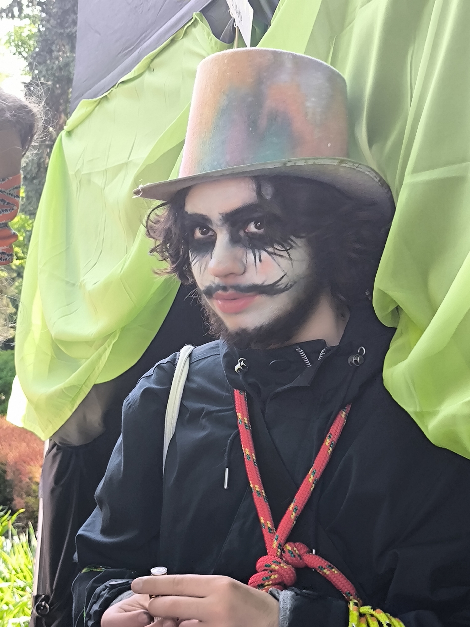 A person wearing face paint and a colorful top hat stands outside, likely at an event, in Riga, Latvia. The person has dark hair, makeup resembling a stylized clown or circus character, and a short beard. They are wearing a black jacket and have a red rope-like necklace or strap around their neck. The background includes green fabric or foliage. In the person's hand, there's a small, unidentifiable object. The setting appears to be in a park or outdoor area with some greenery visible.