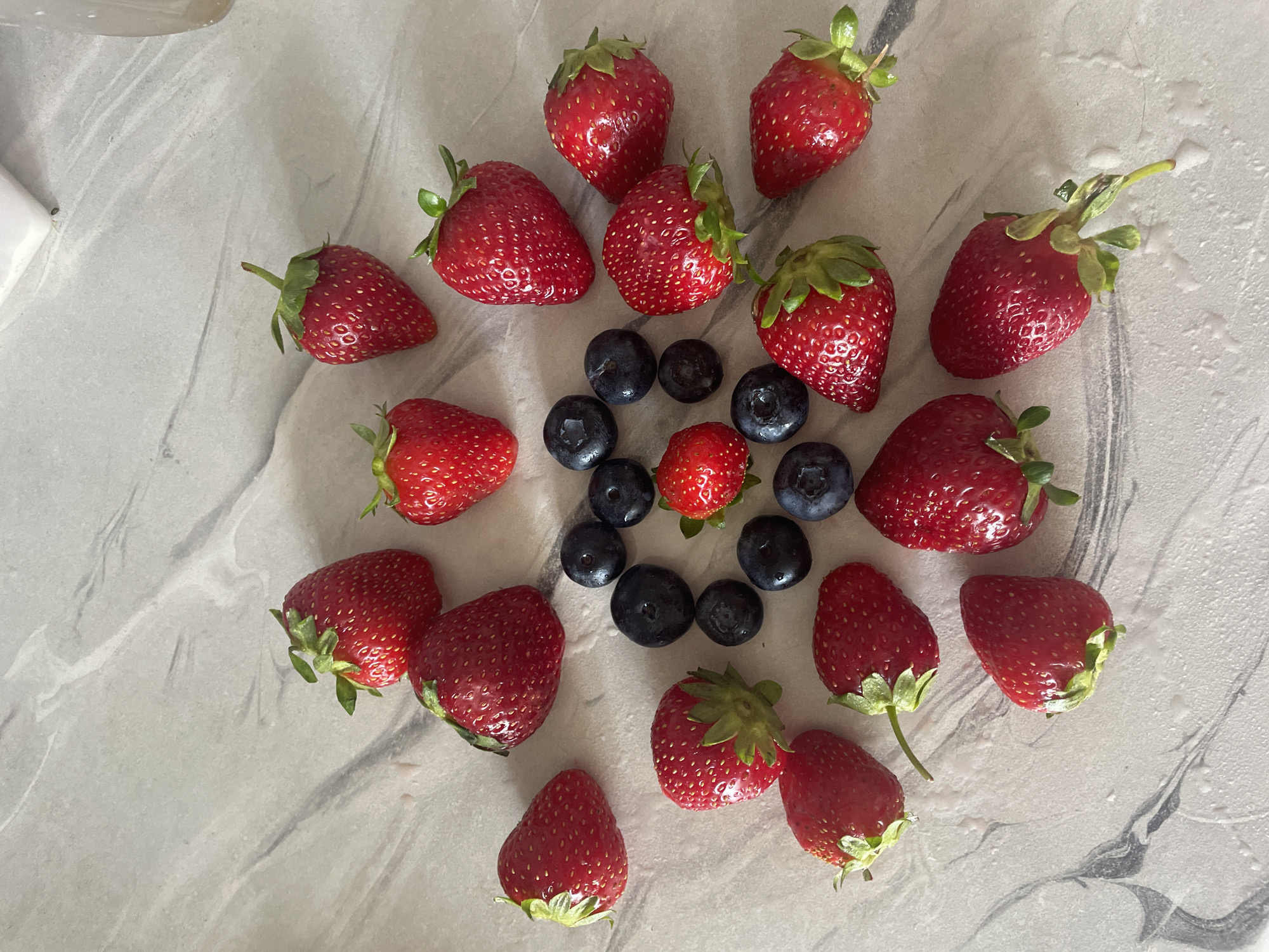 A collection of fresh strawberries and blueberries are arranged in a circular pattern on a light-colored, marbled surface. The strawberries are bright red with green stems and leaves, and some have water droplets on them. The blueberries are dark blue and plump. The arrangement is centered on the surface, with the fruits fanning outwards from the middle. The lighting is natural and bright, casting soft shadows. There are no people or other objects visible in the image. The scene is clean and visually appealing, highlighting the natural beauty of the fruits. There is no information available regarding the location in Brest, Belarus.