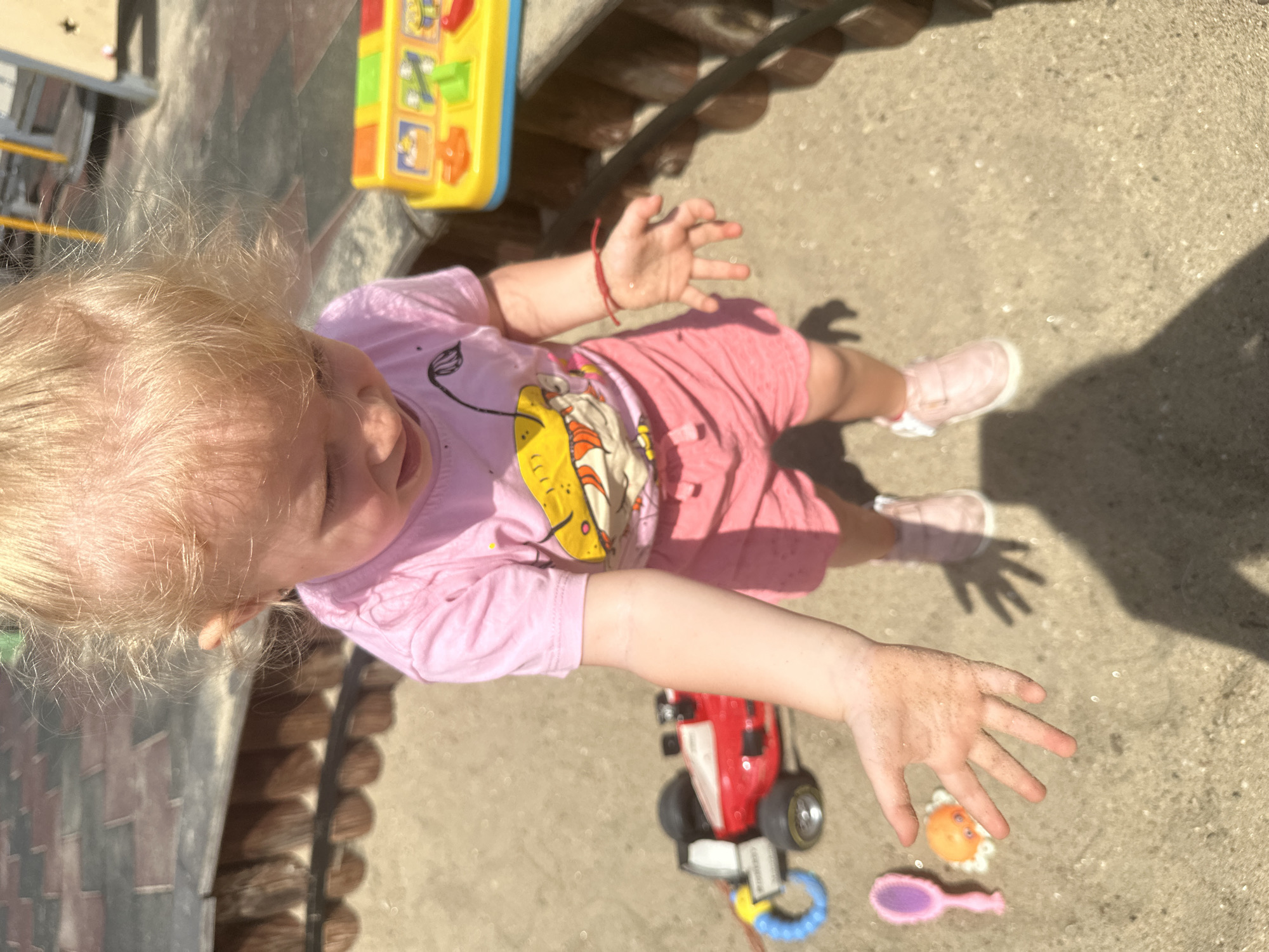 A young child with blonde hair, wearing a pink t-shirt and pink shorts, stands in a sandy area. The child's hands are outstretched and covered in sand. To the left of the child, a red and white toy race car with black wheels and a blue ring is visible. Near the car, a small pink toy brush and a yellow toy with red and green buttons are present. A bright yellow and red plastic toy with various colorful buttons and images is situated to the right of the child, partially obscured by the child's arm. The ground is composed of sand and small pebbles. In the background, a wooden fence-like structure borders the sandy area, with a hint of a playground structure visible further in the distance. The lighting suggests an outdoor, sunny environment. The scene appears to be a playground or sandbox area in Donchovci, Bulgaria.