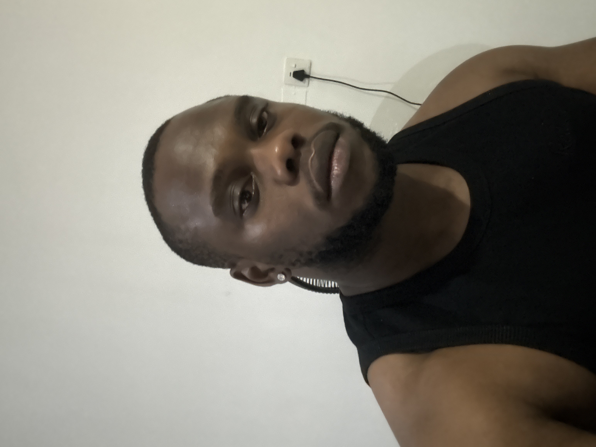 An indoor portrait features a young Black man, likely in his 20s or 30s, captured in a close-up, slightly off-center selfie or photograph. He has a neatly trimmed haircut and a well-maintained beard. He is wearing a plain black sleeveless top or tank top. A small, sparkling stud earring is visible in his left ear. His head is tilted to the right, and he looks directly at the camera with a neutral to slightly serious expression.
The background consists of a plain, light-colored wall, appearing off-white or light gray. On the wall, above and behind his right shoulder, a white electrical wall socket with two slots is visible. A black cable is plugged into the top slot of the socket, extending upwards and out of the frame. The lighting is even, suggesting an indoor setting, possibly with artificial light. There are no other discernible objects or decorations in the background. The scene appears to be simple and uncluttered, focusing primarily on the subject. There is no visible text. The context for the image is Iddo Tudunwada, Nigeria.