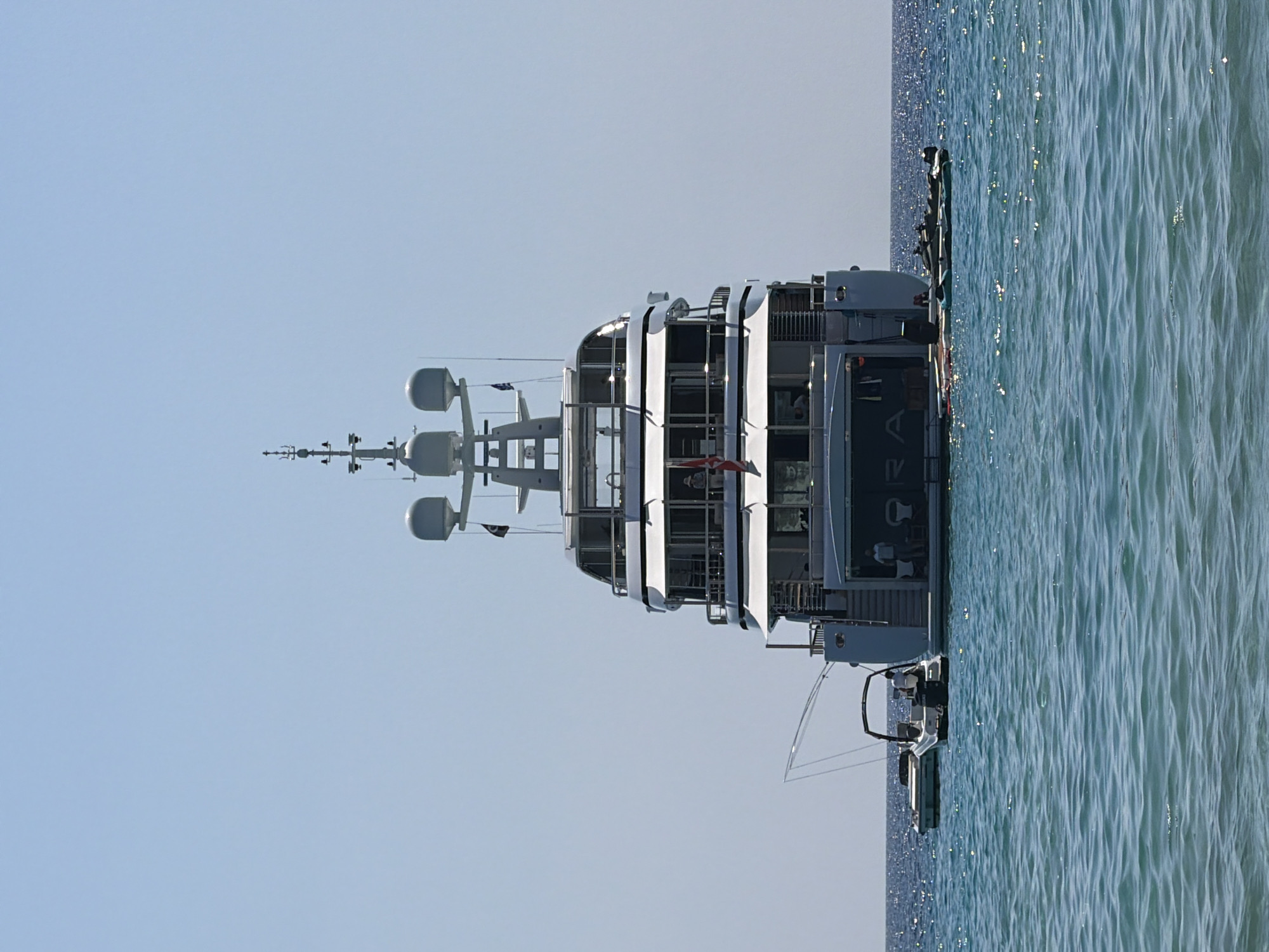 An extreme rear view of a large, white motor yacht is presented, anchored in clear, turquoise ocean water under a bright, cloudless sky. The yacht has multiple decks with windows and balconies. Two smaller boats are positioned near the stern of the yacht, one to the left and another to the right. A person can be seen in the boat on the left, and a few people are visible on the decks and at the open stern of the yacht. The water is calm with gentle ripples and sunlight glinting off the surface. The yacht's name, "ORA," is visible in large letters across its stern.