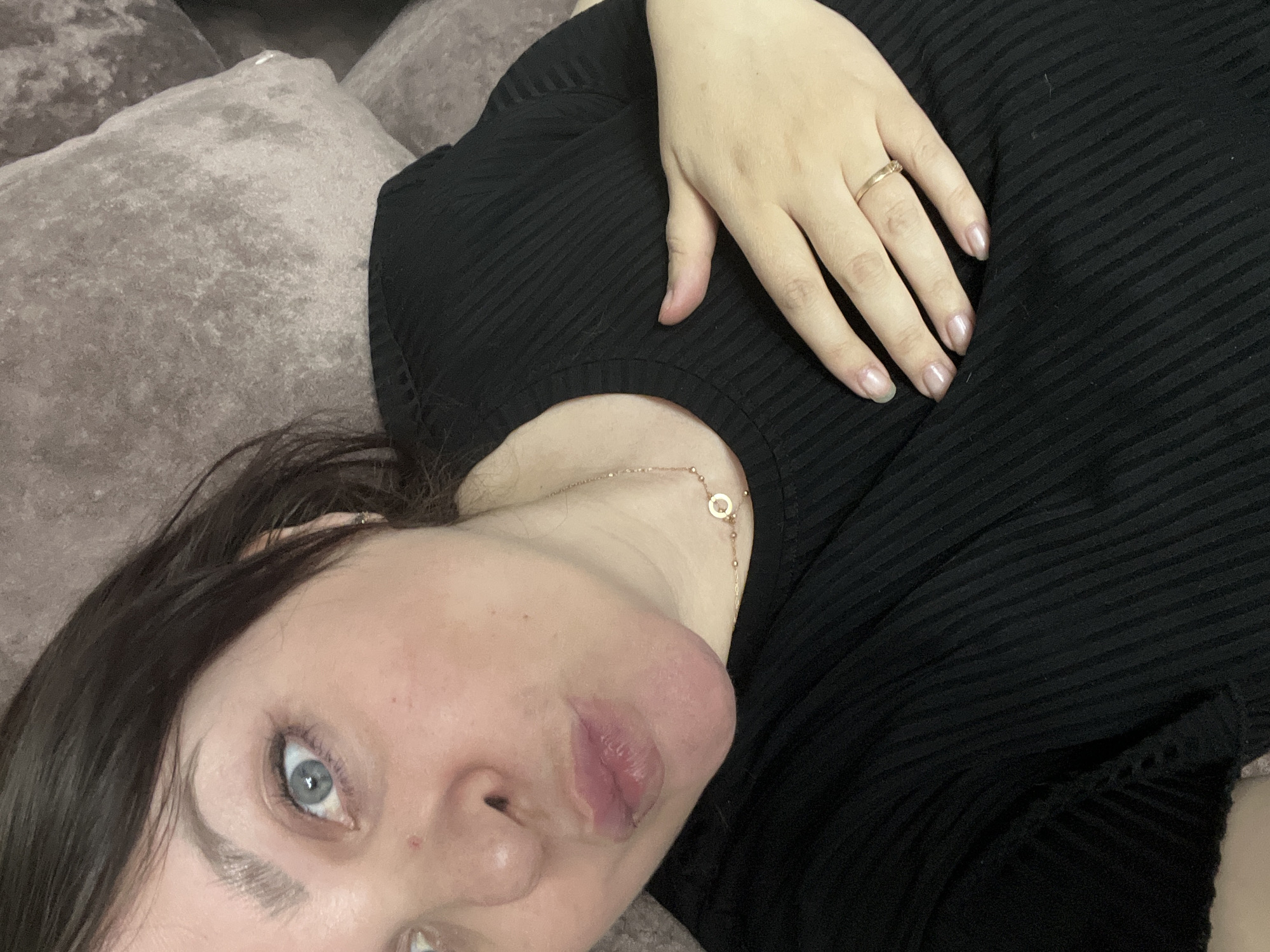 A woman lies on a brown pillow. She wears a black shirt and a gold ring. Her lips are prominent, and she has blue eyes. She wears a gold necklace. The location is unspecified and cannot be determined from the image.