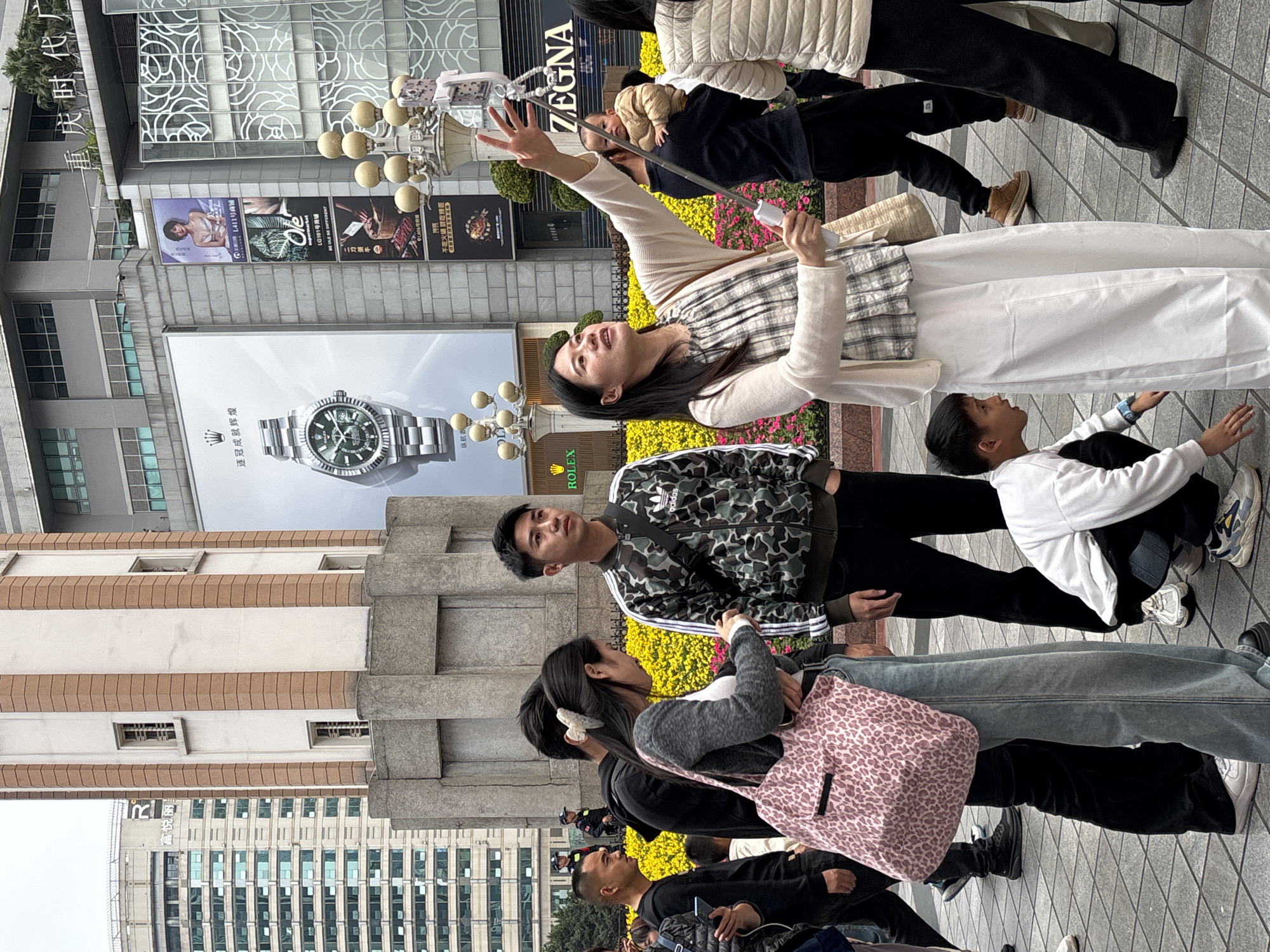 Here is a description of the image.
The image captures a group of people on a city street in what seems to be the Yuzhong District in China. The area is characterized by modern buildings with large storefronts and advertisements. One of the prominent advertisements visible is for Rolex watches.
Several people are present. One woman is taking a selfie with a selfie stick. Another man is wearing an Adidas tracksuit jacket. One person is wearing a jacket. Someone carries a pink, leopard-print bag. There are multiple people standing, walking, or posing in the scene.
The street is filled with blooming yellow flowers adding a pop of color to the urban landscape. The lighting suggests it is daytime.