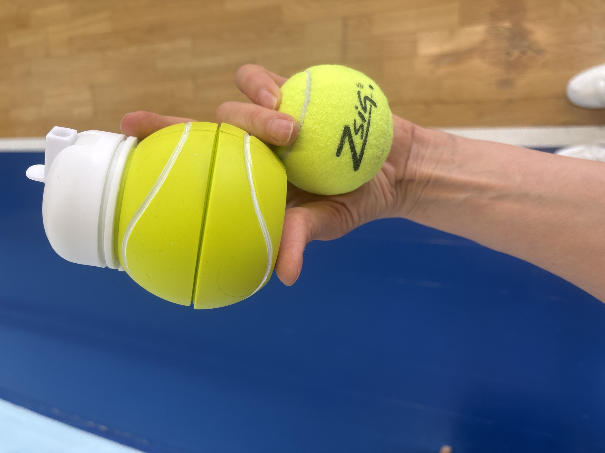 A person holds two yellow tennis balls in their hand. The ball on the left is encased in a white and yellow plastic housing that appears to be segmented. The ball on the right has a black logo on it. The background shows a light brown floor and a blue and light blue surface. The location cannot be determined from the provided image.