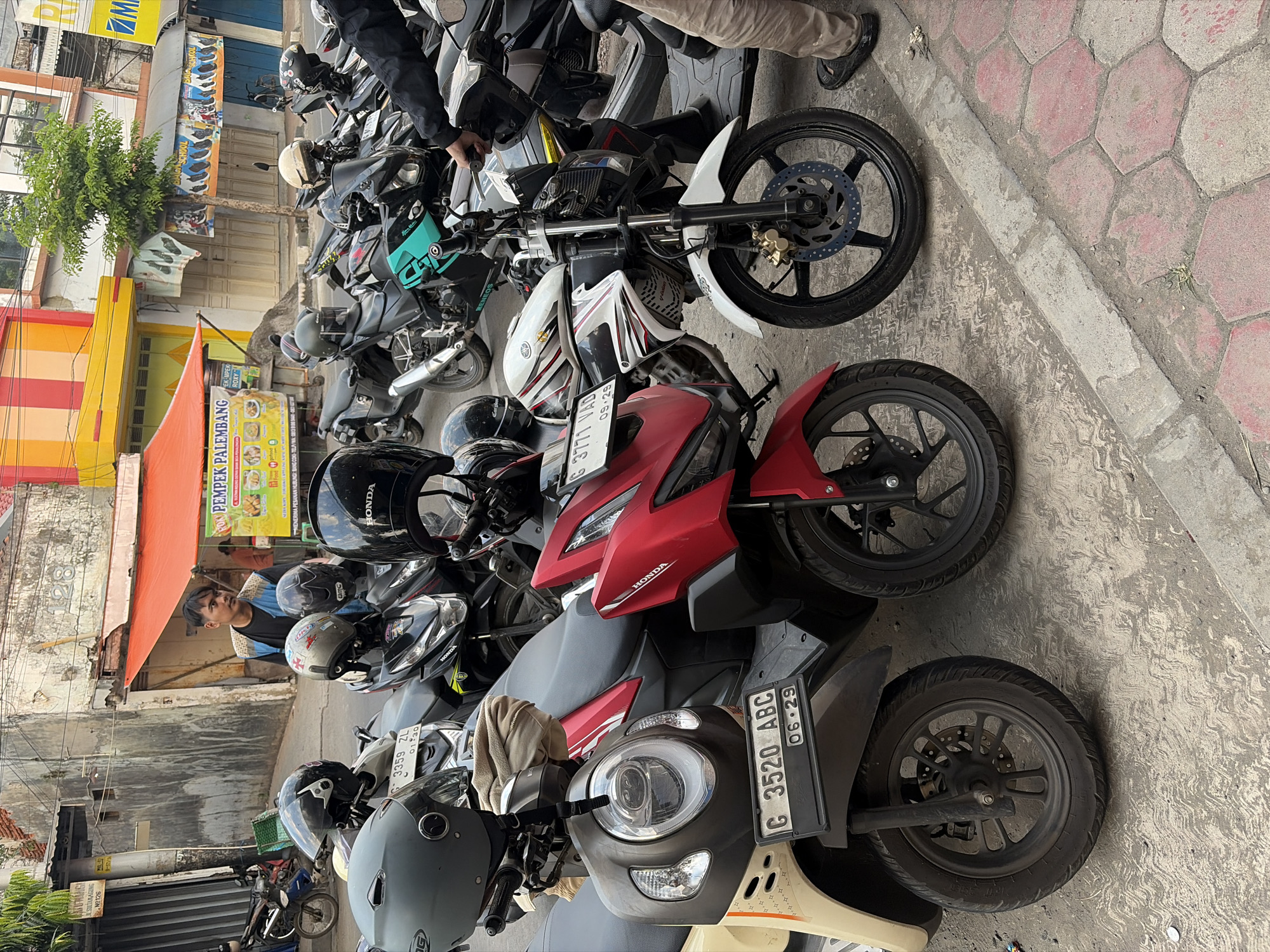 The image depicts a street scene in Pekalongan, Indonesia, with a focus on a row of parked motorcycles. In the foreground, a cream-colored scooter with license plate "G 3520 ABC" is prominent. Next to it, a red motorcycle with license plate "G 3771 VAD" is partially visible. Several other motorcycles, varying in color and style, are parked behind these. Helmets are placed on some of the handlebars. A man wearing a blue vest over a dark shirt and dark pants stands near a food stall that advertises "PEMPEK PALEMBANG." The stall has an orange awning and displays food items. Buildings with weathered facades and colorful awnings line the street. The ground is paved with asphalt and some hexagonal tiles. The overall impression is a typical street view in a bustling Indonesian city, likely during the daytime given the lighting conditions.