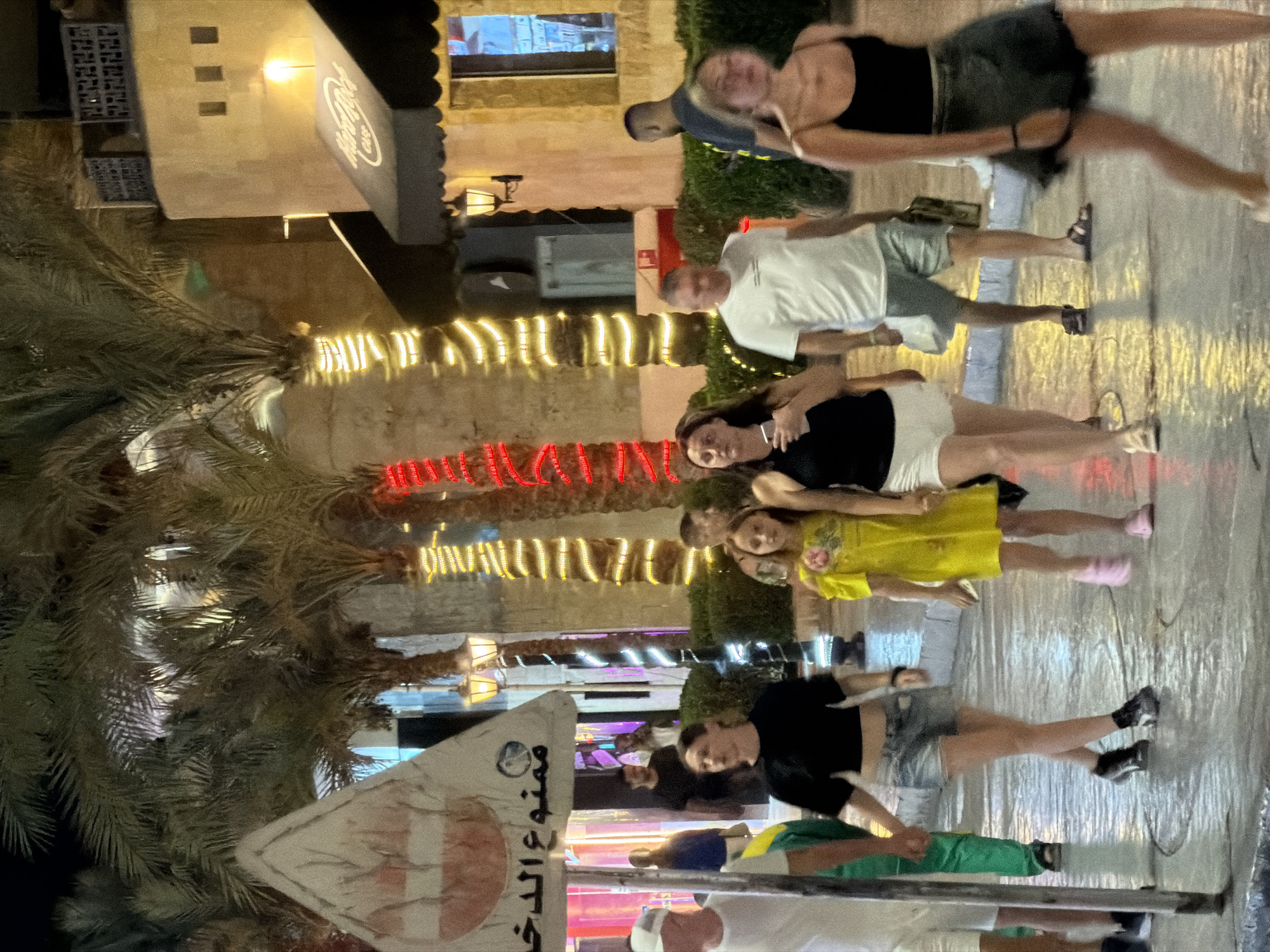 The image depicts a nighttime scene on a street in Sharm El Sheikh, Egypt, possibly within a resort or entertainment district. Several palm trees are visible, adorned with colorful string lights, illuminating the area. A building with a distinctive architectural style, likely a restaurant or shop, is seen in the background, featuring a dark awning with a logo that appears to be for "Hard Rock Cafe."
A group of people are walking on a wet pavement, suggesting recent rain or a cleaning effort. In the foreground, a person wearing a white t-shirt and shorts is walking away from the camera. To their left, a woman in a black crop top and denim shorts is also walking, holding hands with a young girl in a bright yellow dress. Further to the right, another woman in a black tank top and dark skirt is walking towards the camera, with a blurred figure of a man in a white t-shirt and shorts walking behind her. Another man in a dark t-shirt is visible walking further in the background.
A triangular sign with Arabic text and a red circular symbol with a diagonal line through it is present on the left side of the image. The overall atmosphere is lively, with artificial lighting creating a festive ambiance. The wet ground reflects the lights, adding to the visual appeal of the scene.