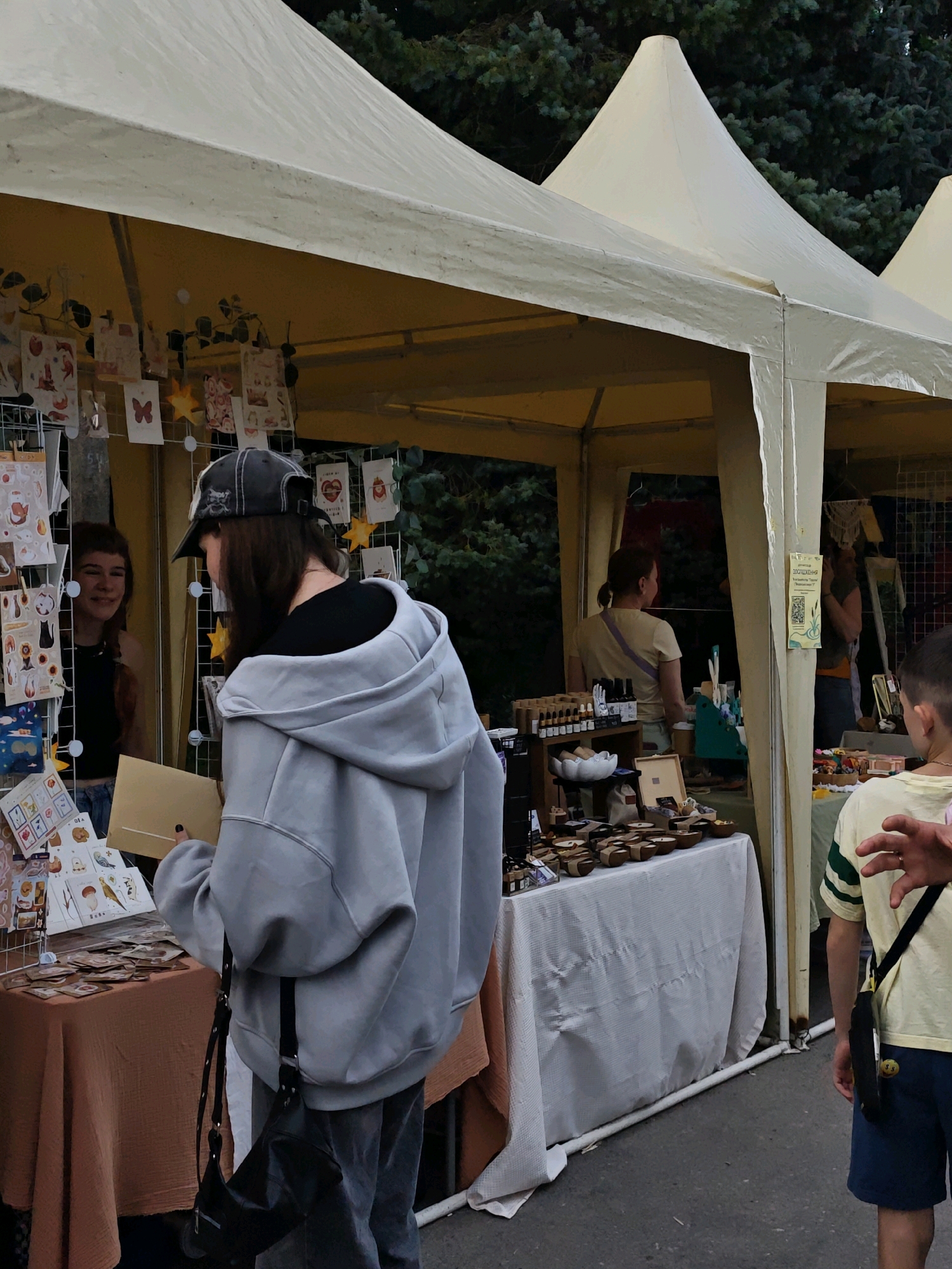 The image depicts an outdoor market scene, likely in Vinnytsia, Ukraine, characterized by tents providing shelter. In the foreground, a person wearing a grey hooded sweatshirt and a baseball cap is examining items at a stall. To their left, another person, appearing to be a vendor, is visible behind a display of small cards and artworks. Further into the market, other stalls are visible with various goods, including what appear to be handmade items and bottled products. The background consists of foliage and trees, suggesting a park or an open urban space. The overall atmosphere suggests a casual, daytime gathering or event.