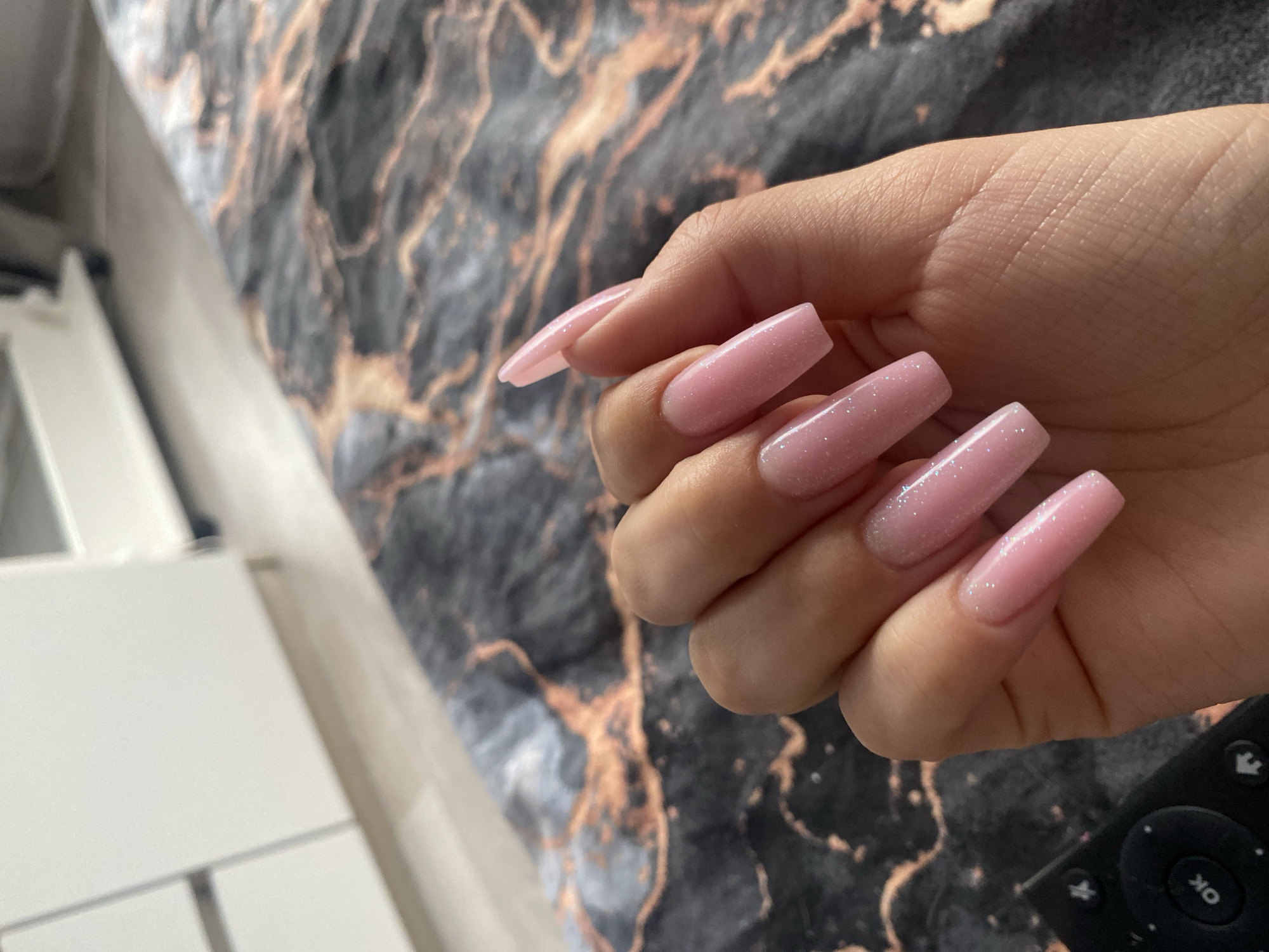 This close-up indoor shot features a right hand displaying long, coffin-shaped artificial nails, painted in a shimmering light pink with fine glitter. The hand is slightly curled, showcasing all five fingernails, which appear freshly manicured. The skin tone is warm, and subtle skin textures are visible.
The hand rests against a dark grey or black fabric adorned with prominent bronze or copper marble-like patterns, likely a bedspread or blanket, which forms a textured background. In the bottom right corner, a portion of a black remote control is visible, featuring several buttons including directional arrows, a central 'OK' button, and a power symbol. To the far left, a blurred light-colored object, possibly a piece of furniture such as a bedside table, is partially visible.
The lighting is bright and even, suggesting it is daytime. The setting appears to be a casual home environment, possibly a bedroom or living room, located in Chaikovsky, Russia. The primary purpose of the image seems to be to showcase the person's detailed and sparkling nails. No specific activity is depicted other than the hand being posed for the photograph.