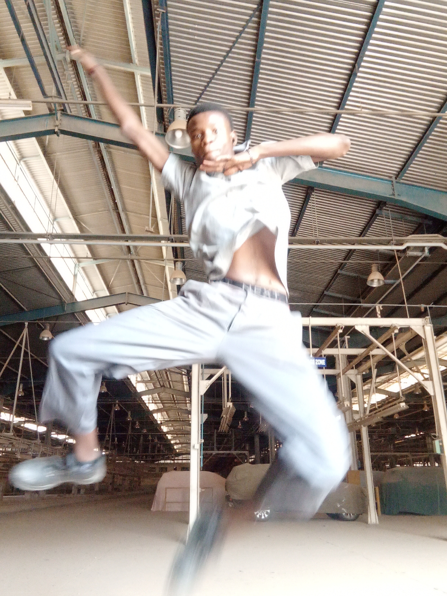 A man is captured mid-air in a blurred photograph, likely jumping. He is wearing a light-colored short-sleeved shirt, light-colored pants, and what appear to be work boots. His right arm is raised above his head, while his left arm is bent at the elbow.
The location appears to be an industrial setting, potentially a factory or warehouse, given the presence of exposed metal beams, a corrugated metal roof with skylights, and hanging light fixtures. There are shrouded objects in the background, suggesting covered machinery or equipment.
The image was likely taken in Kakuri Gwari, Nigeria.