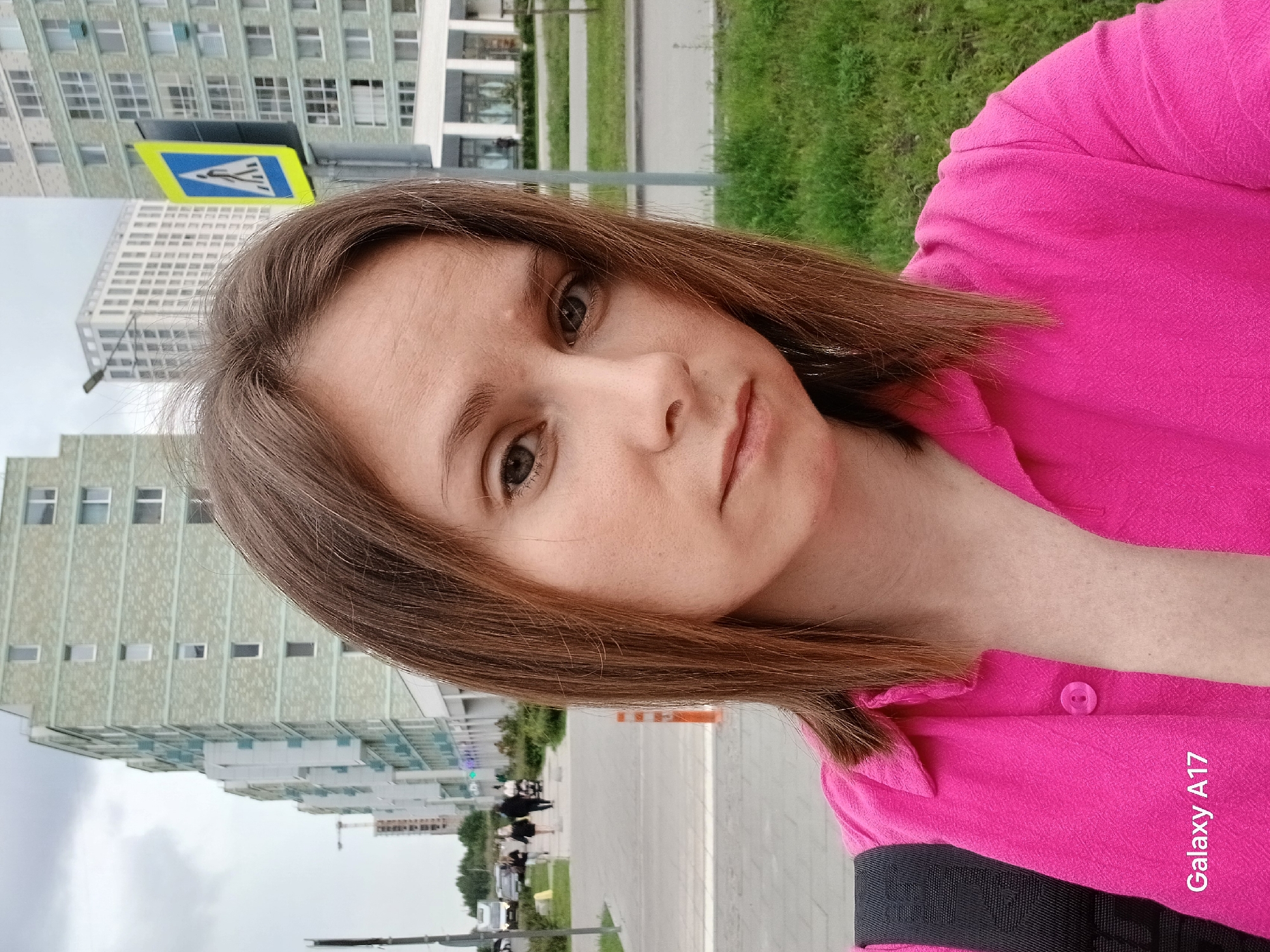 A medium shot selfie is taken from the chest up of a woman with shoulder-length brown hair, looking towards the camera. She is wearing a bright pink button-up shirt. The background shows a street scene in Yekaterinburg, Russia. Behind her are several tall apartment buildings, some under construction and others completed. The buildings are light green and white, with numerous windows. A blue and yellow pedestrian crossing sign is visible on the right side of the image. In the distance, people are walking on a sidewalk, and cars are driving on a road. The ground is paved with grey tiles, and patches of green grass are present. The sky is overcast. The bottom left corner of the image contains the text "Galaxy A17."