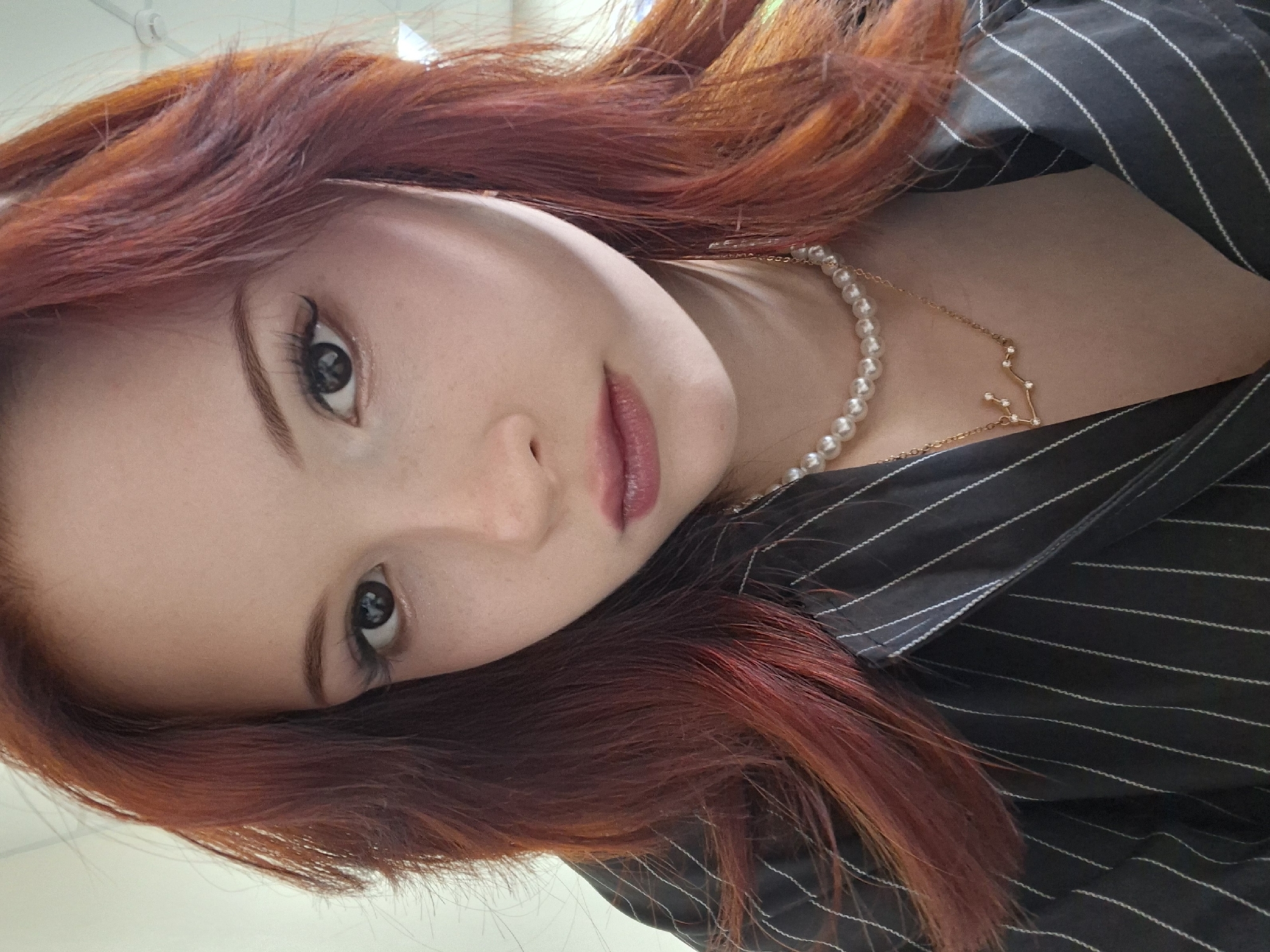 A medium close-up shot depicts a woman with reddish-orange hair, wearing a black and white striped shirt. She is also wearing a pearl necklace and a gold necklace with a constellation design. The setting or location is not discernable from the provided image and cannot be identified as Chaikovsky, Russia or any specific location.