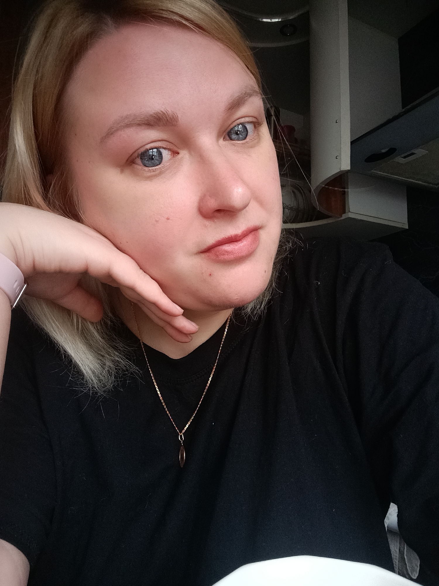 A close-up, chest-up portrait shows a woman with blonde hair and striking blue eyes, captured indoors in Chaikovsky, Russia. She appears to be in her 20s or 30s, with a fair complexion. Her blonde hair is casually styled, with some strands framing her face. She is wearing a simple black t-shirt and a delicate gold-colored chain necklace with a small, elongated pendant. A light pink or beige smartwatch is visible on her left wrist.
The woman is posing for the camera, resting her chin on her left hand, which is gently cupping her jawline. She looks directly into the lens with a neutral to slightly pensive or serene expression. Her lips are closed, adorned with a light-colored lipstick.
The background is dimly lit and largely out of focus, suggesting a domestic interior. Visible elements, particularly on the right side of the frame, include parts of white or light-colored cabinetry, what appears to be a range hood, and a ceiling light fixture, strongly indicating a kitchen setting. The lighting is soft and even, consistent with an indoor daytime environment. The overall mood is casual and intimate. No text is visible in the image.