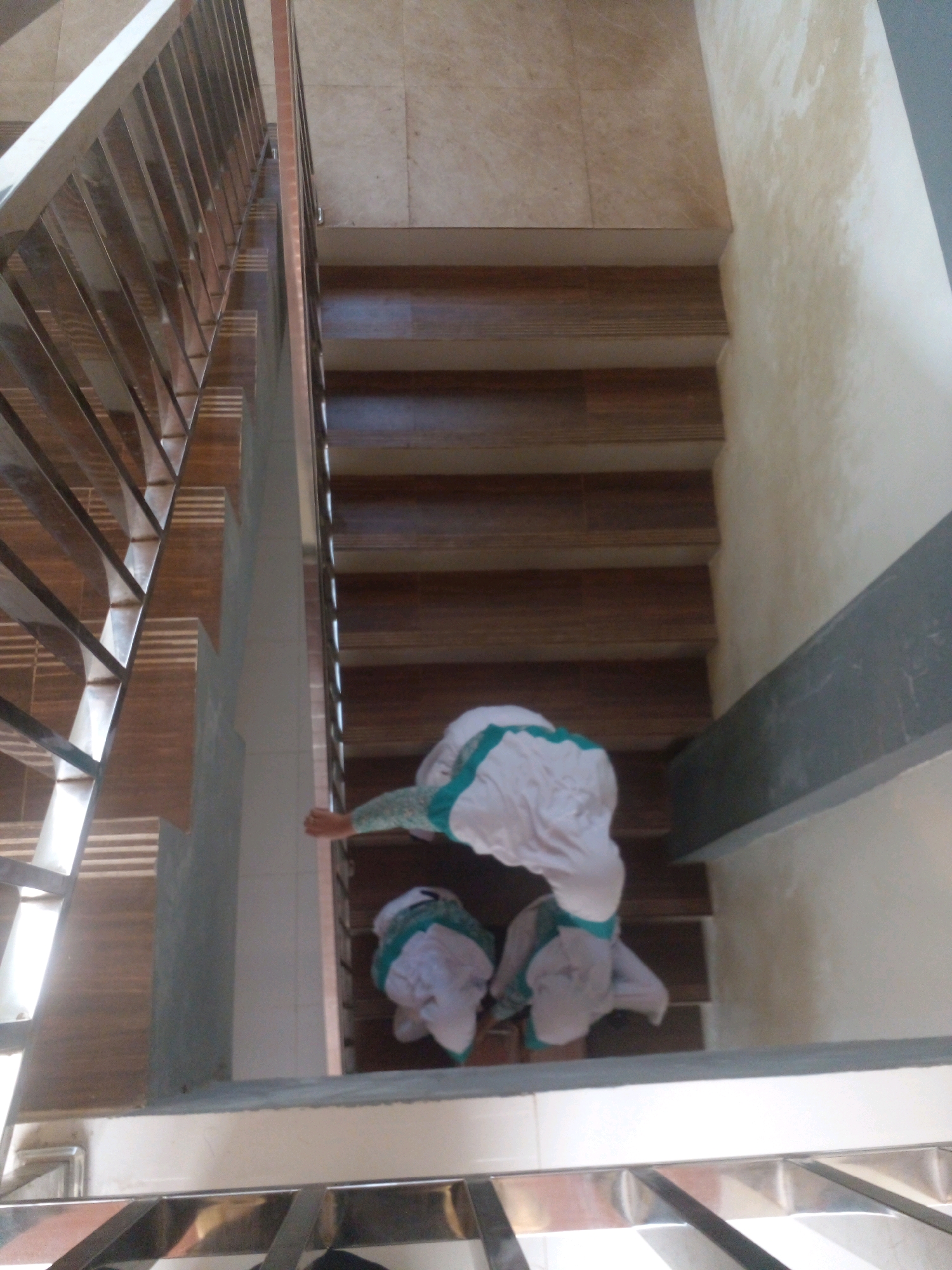 In the city of Banyuputih, Indonesia, a high-angle shot captures a staircase with two individuals descending. The staircase features polished wooden steps and a metallic railing on the left. The walls are tiled with a light beige color, and a concrete beam runs across the right side of the frame. The first individual, positioned higher on the stairs, is wearing a white garment with green trim. They are actively descending, with their right hand extended slightly downwards, possibly reaching for the railing. Below them, a second individual, also in a white garment with green trim, is positioned lower on the stairs, appearing to be moving downwards as well. Both individuals' faces are not visible, and their clothing suggests they may be wearing hijabs. The overall scene is captured from above, looking down the staircase.