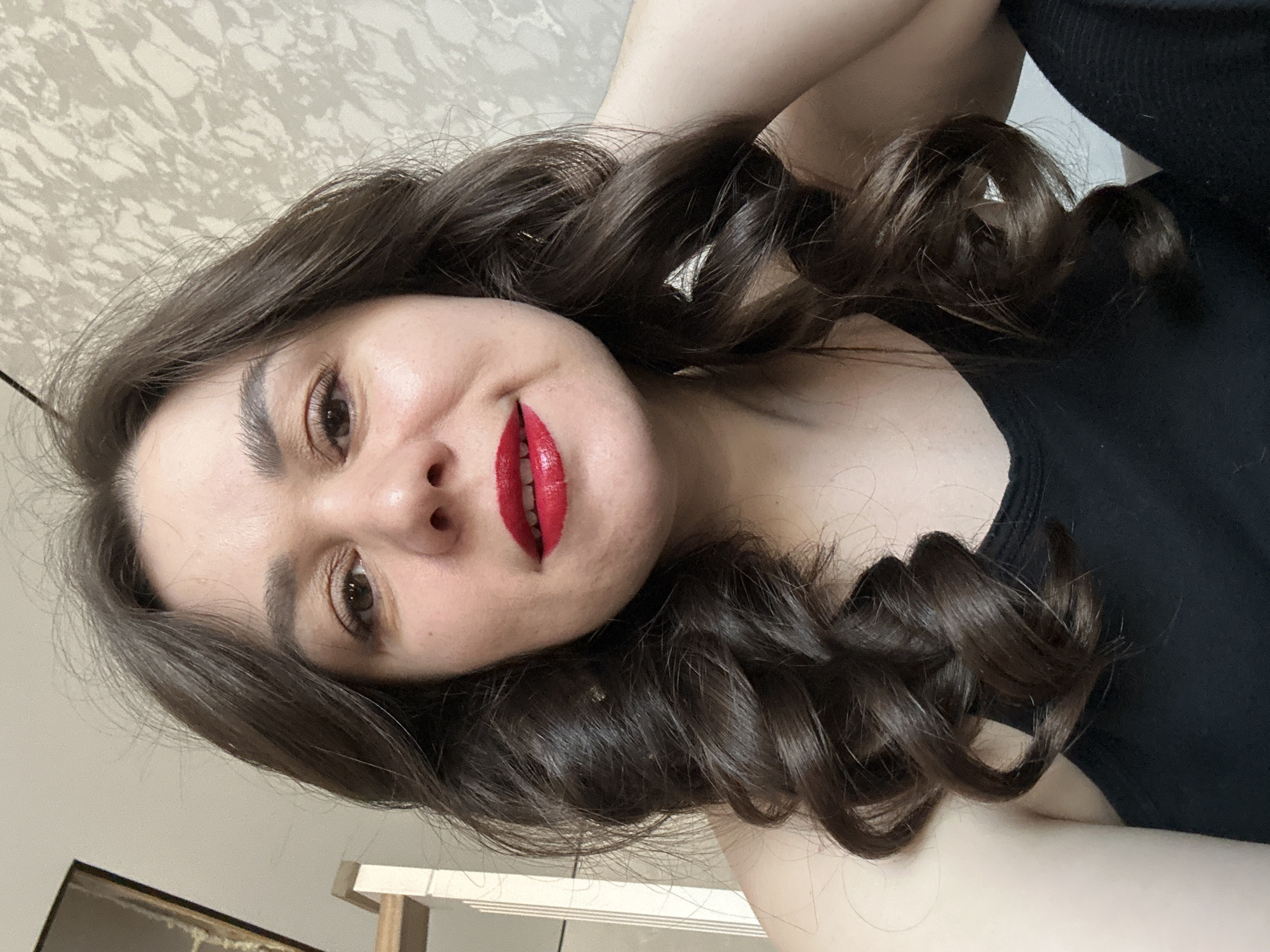 A woman with fair skin, dark brown hair styled in loose curls, and red lipstick poses for a selfie. She is wearing a black tank top. The background includes a patterned wall and a glimpse of a room with a light-colored wall and some furniture. The photo may have been taken indoors, but no specific location in the city of Chaikovsky, Russia is identifiable.