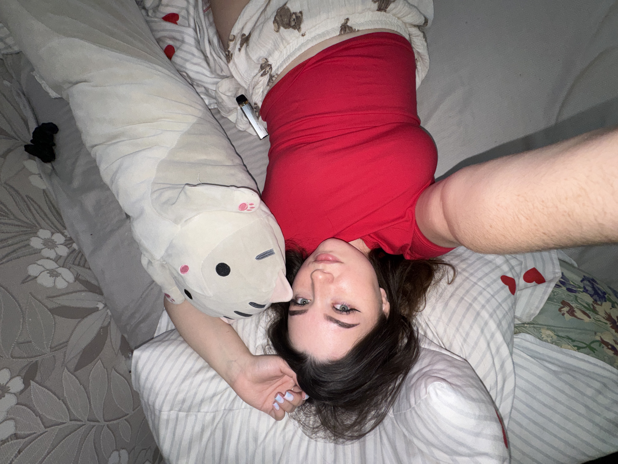 A young woman is depicted lying on her back on a bed, looking directly at the camera. She has dark hair, light-colored eyes, and well-defined eyebrows. She wears a vibrant red sleeveless top and white pajama shorts adorned with small brown bear patterns. Her fingernails are painted a pale blue.
Beside her head is a large, light-colored body pillow shaped like a cartoon cat or animal, featuring black circular eyes and pink ear details. A metallic silver vape pen is visible, tucked into the waistband of her shorts. The bed is covered with an assortment of bedding, including a grey and white floral patterned sheet to her left, a striped sheet beneath her, and a green floral sheet with botanical patterns on the right. Another striped pillow with red heart decals is visible behind her. A small, crumpled black item, possibly clothing, lies on the patterned sheet to the left. The scene suggests a relaxed moment in a bedroom setting, likely captured as a casual selfie or portrait. The location context for this image is Башкултаево, Russia.