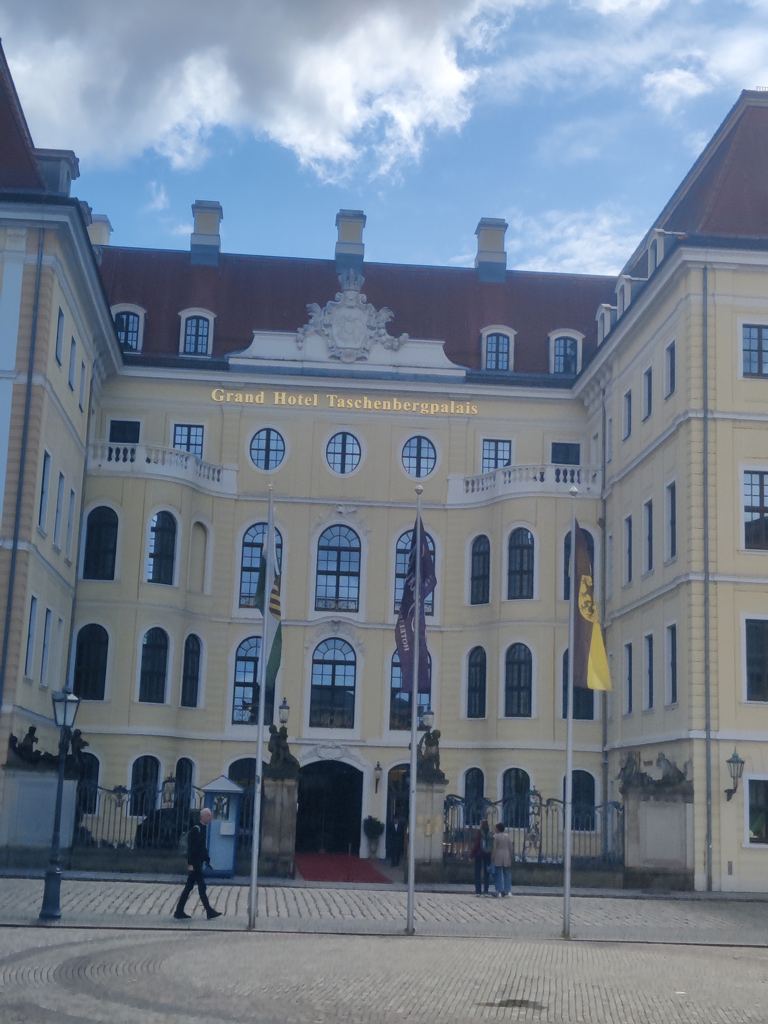The image captures the exterior of the Grand Hotel Taschenbergpalais in Dresden, Germany, under a partly cloudy sky. The imposing building features a pale yellow facade with a red tiled roof and numerous windows of various shapes and sizes. The "Grand Hotel Taschenbergpalais" is prominently displayed in golden lettering above the entrance.
In the foreground, a person in a dark suit is walking across a cobblestone street. Further back, near the entrance, two individuals stand by a fence, one wearing a brown jacket and the other a blue jacket. Flags, one purple and one yellow, are flown from poles in front of the hotel. The architecture suggests a historical and grand setting, typical of Dresden's city center. The lighting indicates it is daytime, with shadows cast by the building and people.