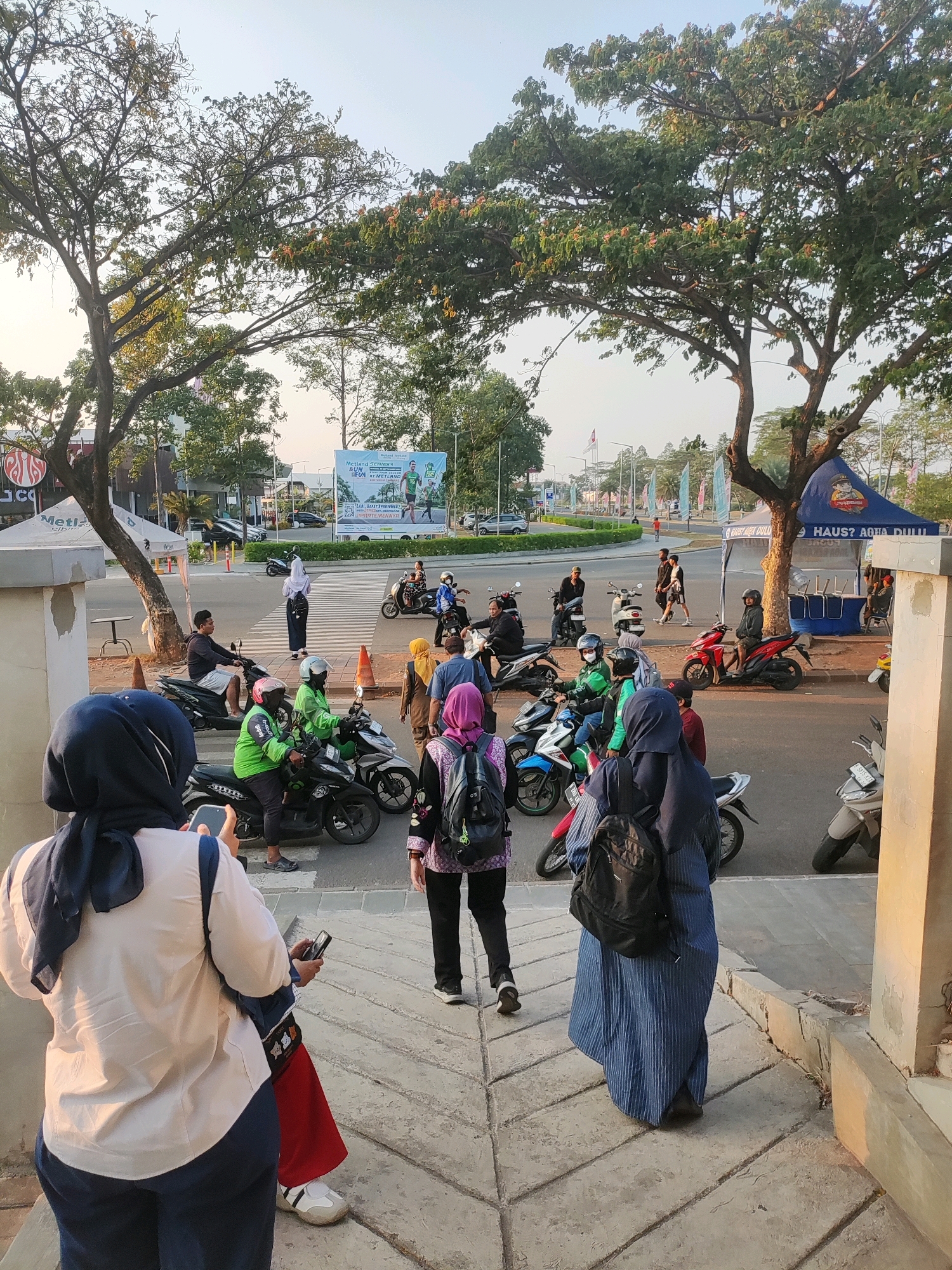 The image depicts a street scene in Telagamurni, Indonesia, with a clear focus on a pedestrian crossing. Several people are visible, many of whom are wearing hijabs. Some are standing, while others are on or near motorcycles. In the foreground, two women in hijabs are walking away from the camera, with one holding a smartphone.
The street is lined with trees, and there is a large banner in the background advertising a "Melang Series Run" event. A blue tent with "HAUS? AROMA DULU" written on it is set up to the right of the frame. Several motorcycles, some parked and some being ridden, are present on the road and sidewalk. The overall atmosphere appears to be that of a busy urban environment, possibly during an event or a typical day with a significant amount of motorcycle traffic. The lighting suggests it is either early morning or late afternoon, with the sun casting long shadows.