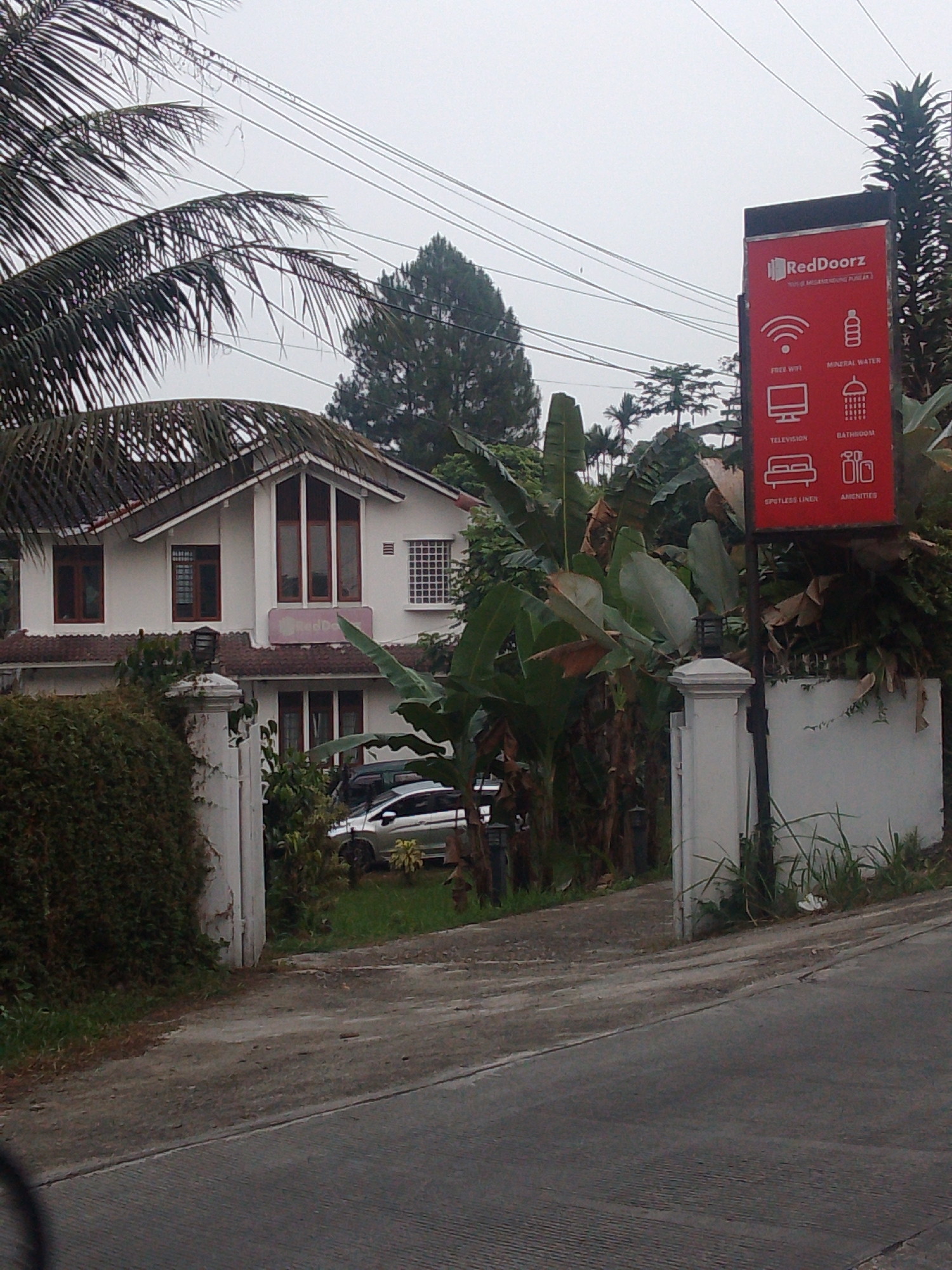 The image shows a white two-story house with a red roof, identified as RedDoorz in Megamendung, Indonesia. A prominent red sign displaying the RedDoorz logo and amenities such as free WiFi, mineral water, television, bathroom, spotless linen, and amenities is visible. The house is surrounded by lush greenery, including palm trees and banana plants. A paved driveway leads to the entrance, where two cars are parked. The sky is overcast, suggesting a cloudy day. The overall scene depicts a tranquil setting in a residential area, likely a hotel or guesthouse.