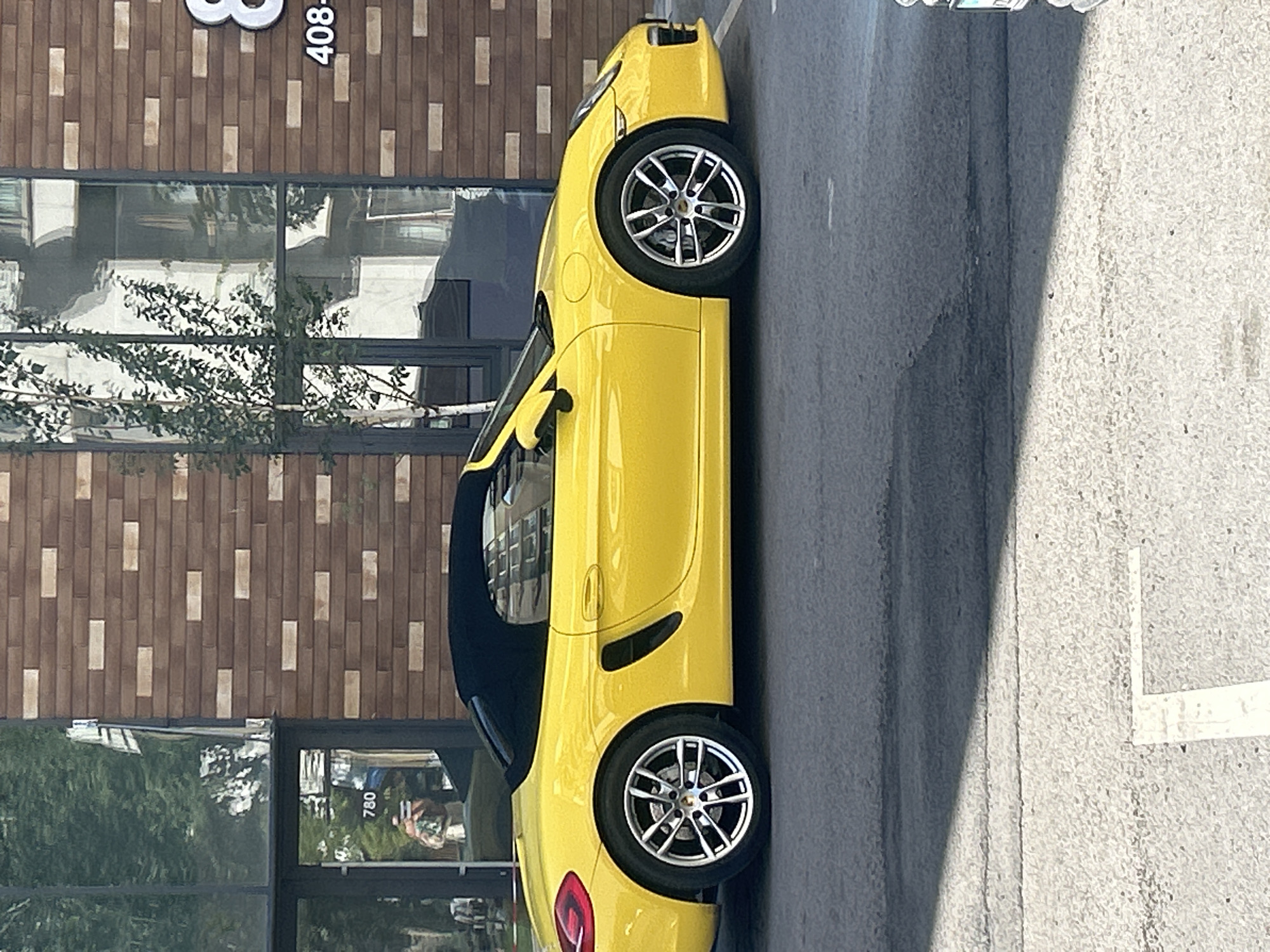 A bright yellow convertible Porsche is parked in front of a building with a brick facade. The building has large windows that reflect trees and other parts of the city. A reflection in the glass of the building shows a person holding a phone. The building number "780" is visible on a glass door. The number "8" and "408" are visible on the brick facade. The scene appears to be a street or parking lot in Almaty, Kazakhstan.