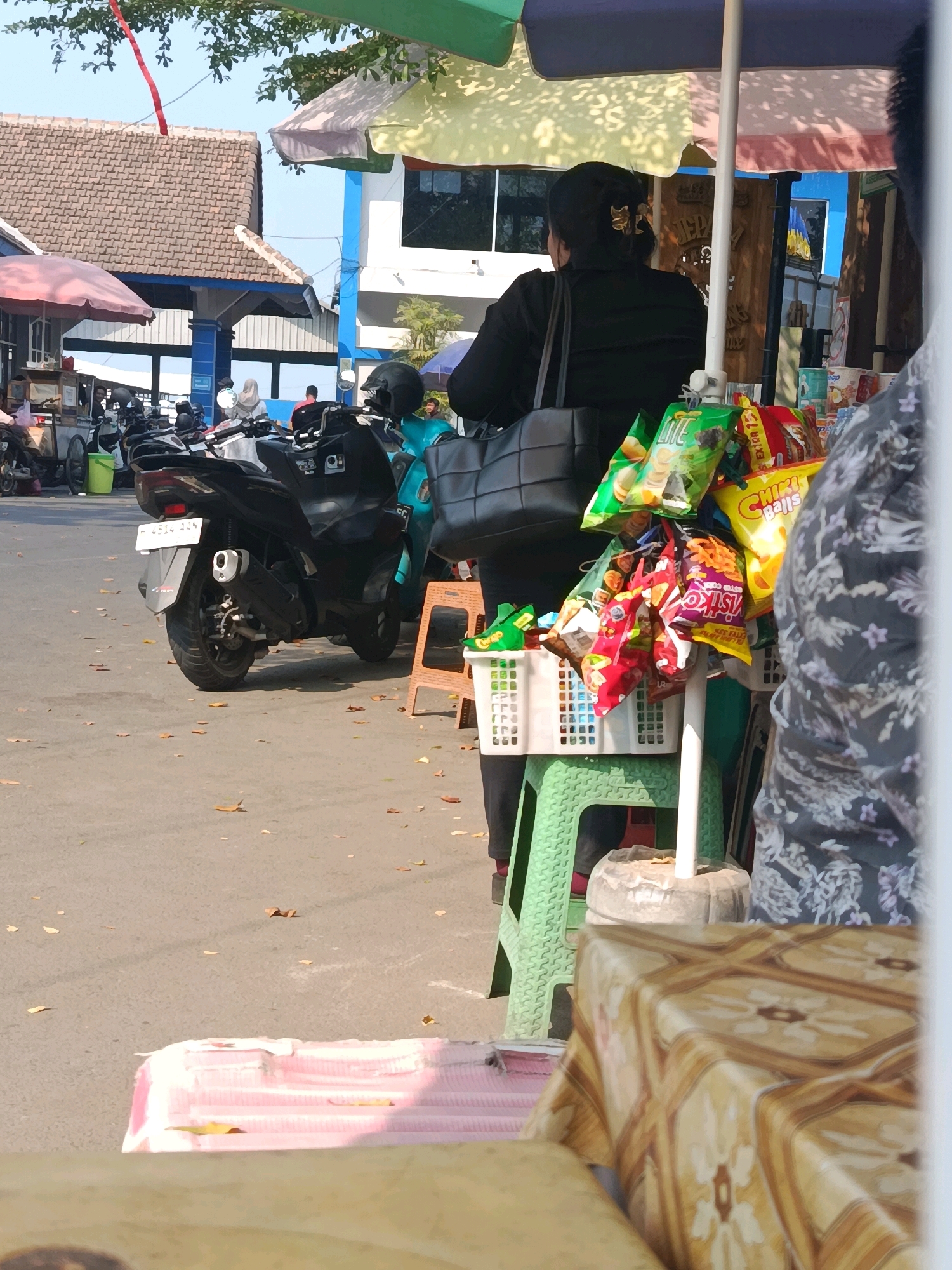 A woman wearing a black shirt and black pants stands behind a display of snacks under a multi-colored umbrella. The snacks are in various colorful bags and a white basket. A motorcycle is parked to her left, and other parked motorcycles and a cart are visible in the background. Buildings with tiled roofs and blue accents are also in the background. The scene appears to be outdoors on a paved surface with fallen leaves scattered around. A patterned tablecloth is visible in the foreground. The signage in the background reads "JEPARA".