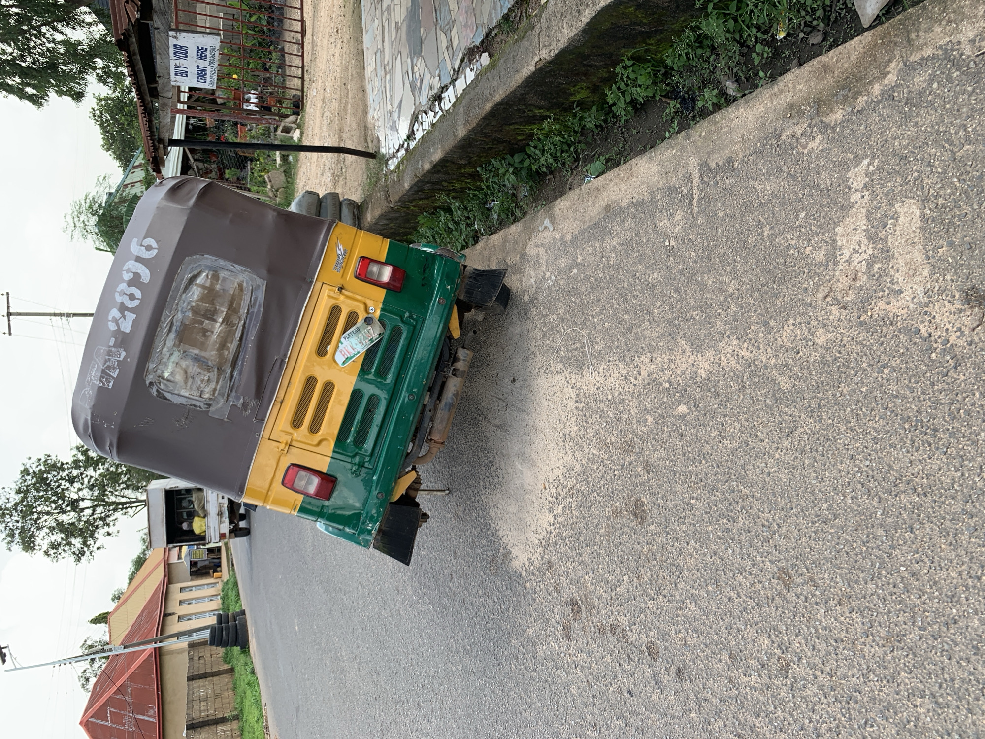 An auto-rickshaw, painted green and yellow with a brown canopy, has overturned onto its side on a paved road. The auto-rickshaw's license plate reads "PLATEAU". The vehicle is angled precariously, with its rear wheels in the air. The road appears to be in a commercial area, as indicated by a "BUY YOUR CEMENT HERE" sign visible on a gate in the background. Buildings with red-tiled roofs are visible behind a brick wall to the left. The environment suggests it is daytime, with overcast skies. The scene appears to be located in Jos, Nigeria.