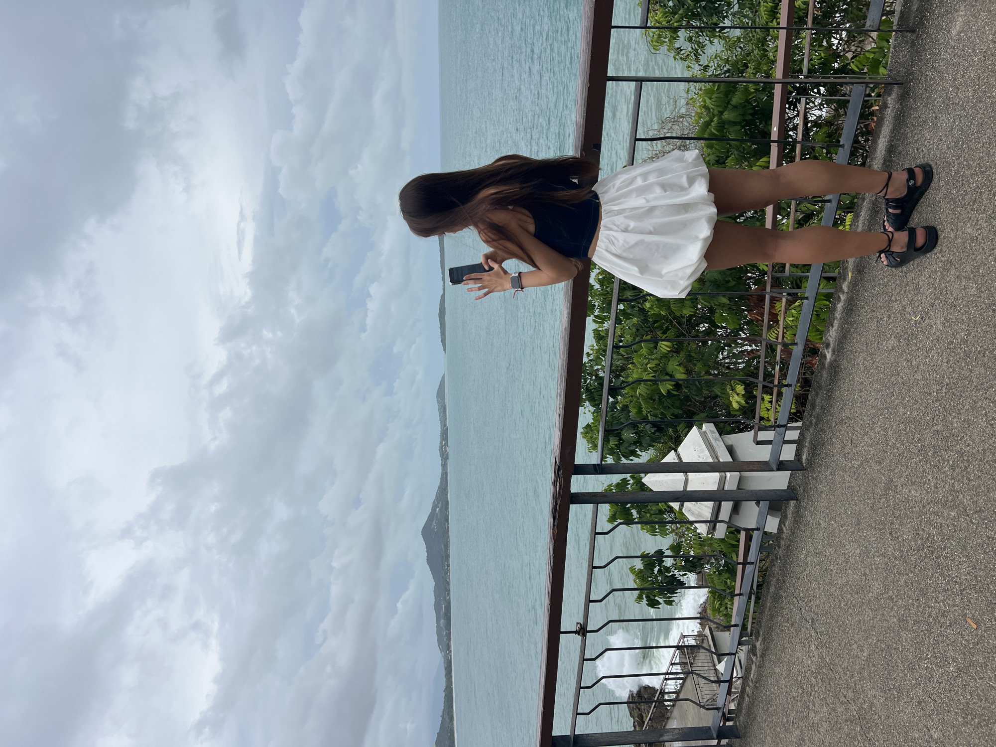 A young woman with long brown hair is standing on a concrete patio behind a black wrought-iron railing, overlooking the ocean, presumably at the city of Baan Chaweng Noi, Thailand. She is holding a smartphone in her hands, appearing to take a picture or video. She is wearing a navy blue top, a short, white skirt, and black sandals with ankle straps. The sky is overcast with grey clouds, and the water is a pale green-blue color. In the distance, a land mass is visible in the ocean. Green foliage is growing near the railing. The patio is slightly tilted, with a building or structure with white elements visible in the lower right corner.