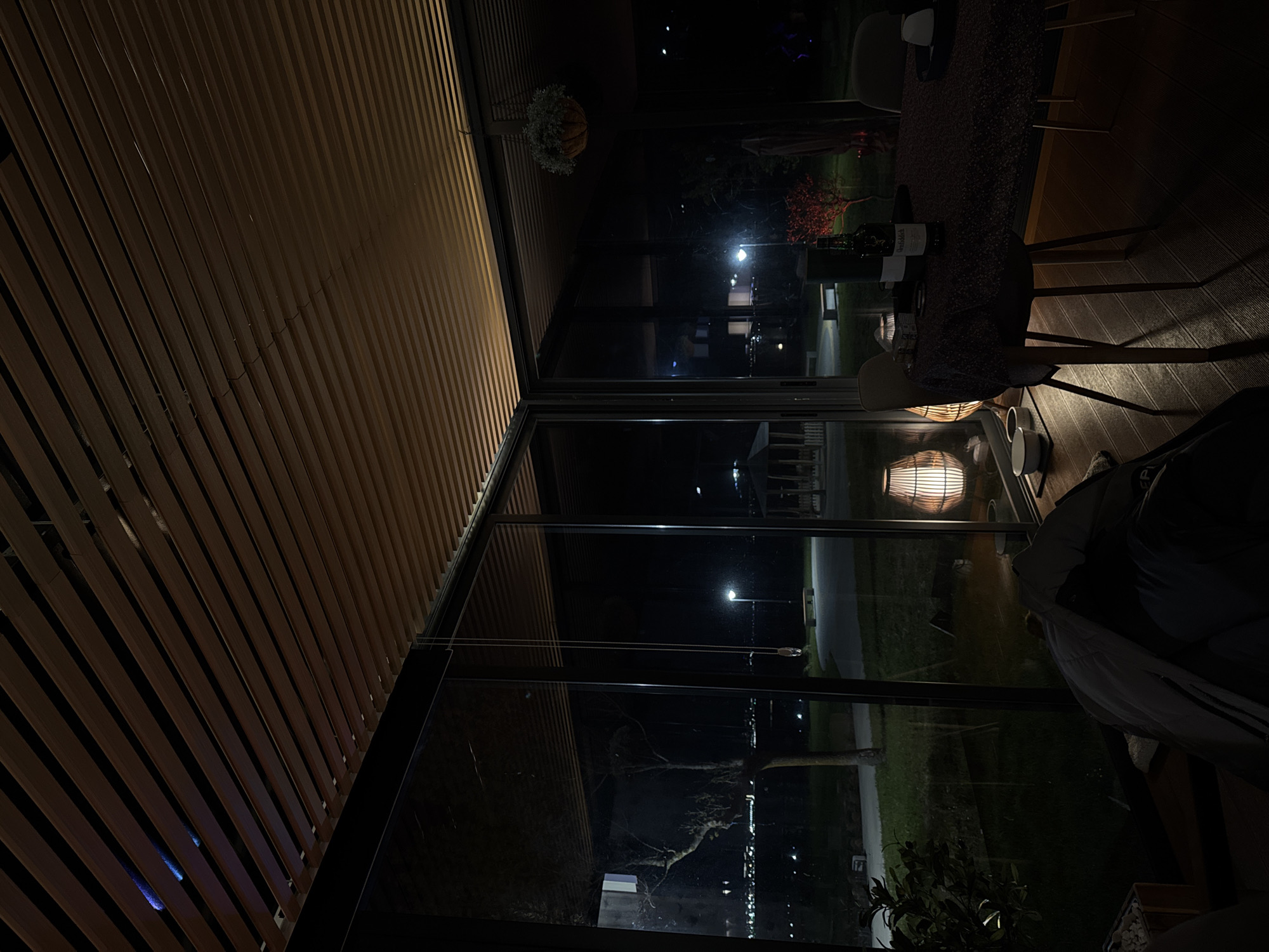 The image depicts an interior space with a view of a night scene in Burgas, Bulgaria. The foreground consists of a wooden-slatted ceiling and large glass windows that look out onto a cityscape. In the interior space, there are dining tables with chairs, indicating a restaurant or dining area. On one of the tables, there are bottles of wine and possibly wine glasses. A person is seated, partially visible, with a light-colored jacket.
Outside the windows, there is a street with streetlights, trees, and buildings in the background. The lights suggest it is nighttime. The reflection in the glass adds complexity to the view, making it difficult to precisely discern the exterior environment.