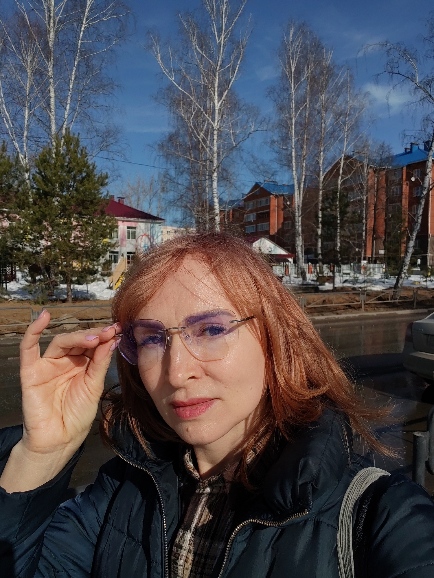 A self-portrait captured outdoors in Chaikovsky, Russia, features an adult female under a clear blue sky. She possesses fair skin and reddish-brown hair, holding light-framed eyeglasses with purple-tinted lenses by the right temple with her left hand, which exhibits light pink nail polish. She is wearing a dark blue puffer jacket over a plaid shirt. The background includes bare birch trees, green coniferous trees, and multi-story brick residential buildings topped with blue roofs. A smaller building with a red roof and playground equipment is also present. Portions of a paved road, a low metal fence, and patches of snow on the ground indicate a late winter or early spring environment. A light-colored vehicle is partially visible on the far right.
