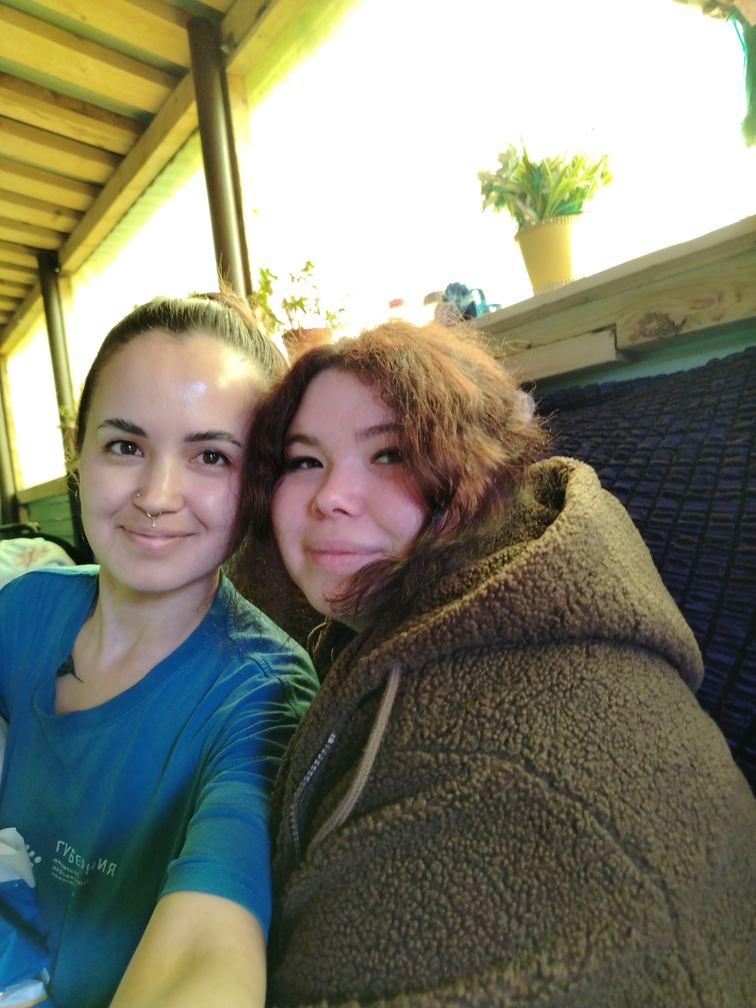 Two women are posing for a selfie indoors. The woman on the left has her hair pulled back in a bun, and wears a blue t-shirt. The woman on the right has auburn hair and wears a brown, sherpa-style hooded sweatshirt. The location is Гожан, Russia. Behind them is a windowsill with a potted plant. The area appears to be a porch or covered outdoor space due to the wooden beams in the background.