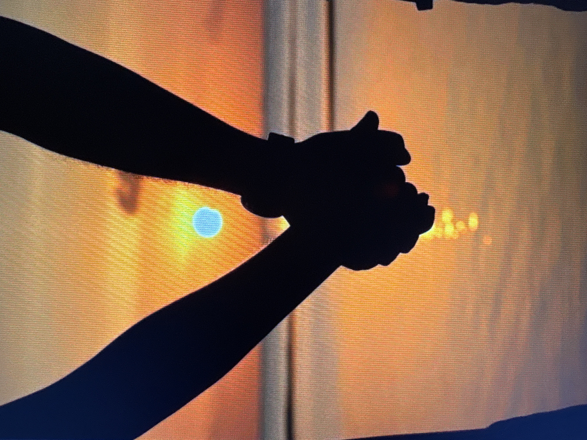 The image shows a silhouette of two people holding hands. They are standing on a beach with the ocean and a sunset in the background. The sun is a bright orange disk with rays extending outwards. The sky is a gradient of orange, yellow, and blue. The water reflects the colors of the sunset, creating a shimmering effect. The silhouettes of the people are dark and indistinct, making it impossible to determine their features or clothing. The image is framed tightly on the hands and arms of the couple. The image is taken from a low angle, looking up at the couple and the sunset. There is no indication of the city of Kuvshinovo, Russia in the image or any context to place it there.