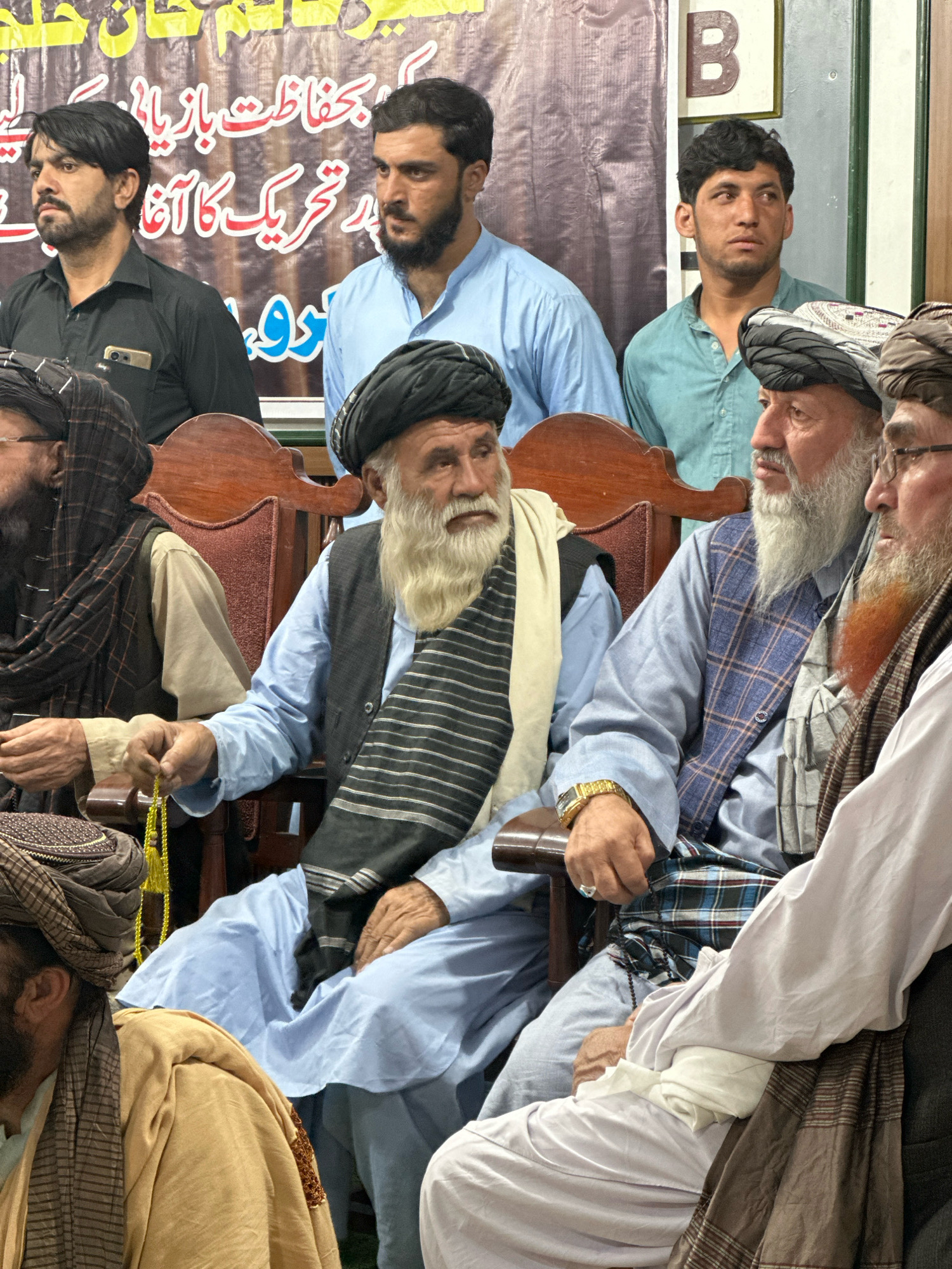 In Quetta City Tehsil, Pakistan, a group of men, predominantly wearing traditional Islamic attire, are gathered indoors. The setting appears to be a formal event or meeting, with men seated on wooden chairs in front of a backdrop featuring text in an Urdu script. The men are diverse in age, with some sporting beards and turbans. One man in the foreground, holding a string of yellow beads, is wearing a light blue shirt and a dark vest adorned with a striped sash. Another man beside him wears a blue plaid vest over a light blue shirt. The environment is lit, suggesting an indoor space illuminated by artificial lighting. The overall atmosphere is one of solemnity or respectful attendance.