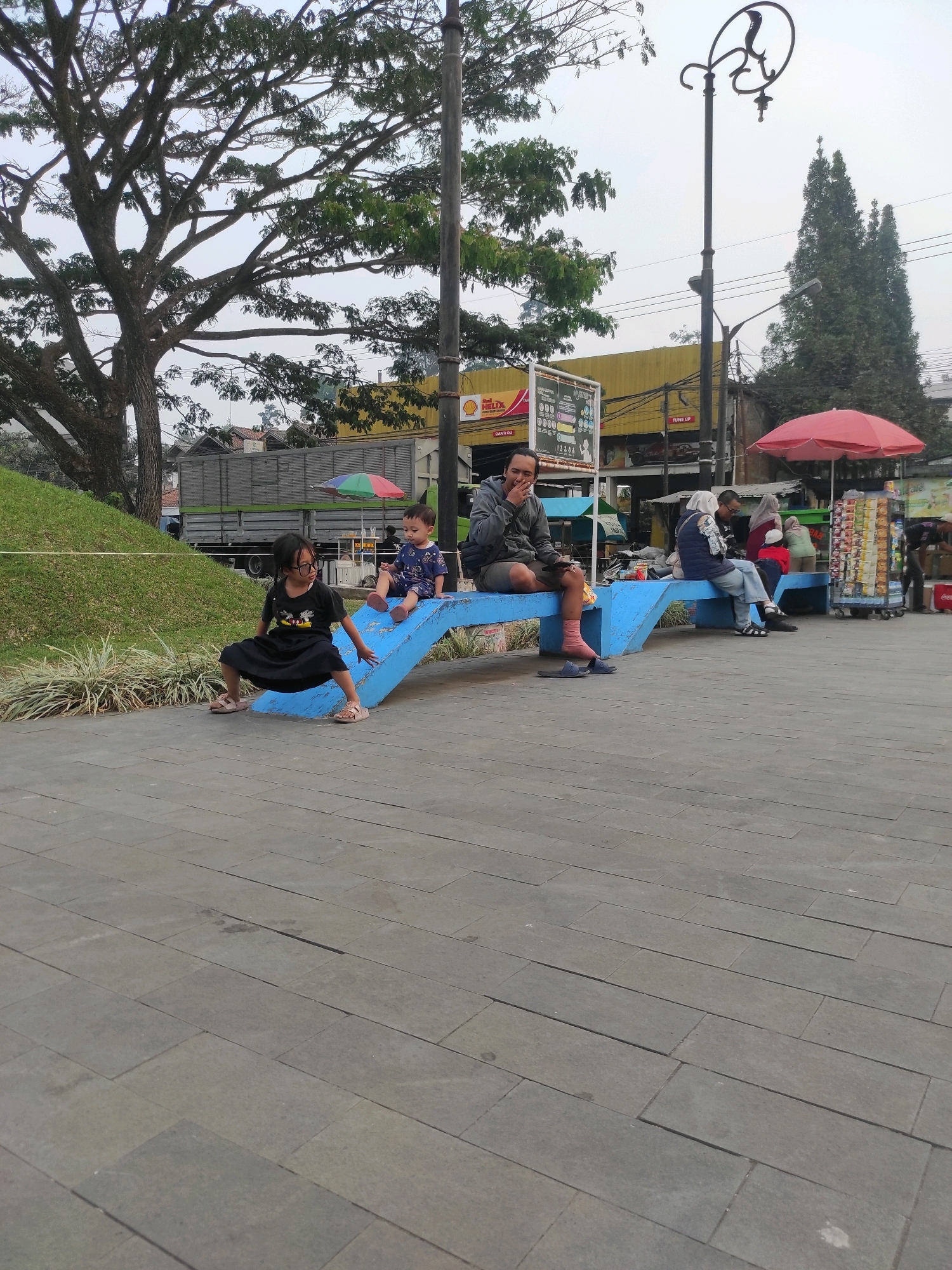 In Padalarang, Indonesia, a man sits on a blue bench with his hand over his mouth, appearing to laugh or cough. To his right, three people are seated on the same bench; one woman wears a headscarf and blue vest, another woman wears a red headscarf, and a child is seated on her lap. Further to the right, a vendor stands behind a cart filled with packaged goods. In the foreground, a young girl in a black dress and glasses crouches on the blue bench. To her right, a toddler in a blue shirt and shorts sits on the bench. The scene takes place on a paved walkway, with a grassy hill and trees in the background, along with a truck and commercial buildings. A decorative lamppost with a snake motif stands near the center of the image.