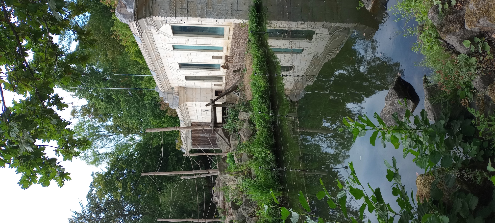 The image shows an outdoor enclosure at the Zoom Erlebniswelt Gelsenkirchen, a zoo in Gelsenkirchen, Germany. The enclosure features a rocky landscape with several wooden poles and ropes, suggesting it is designed for primates. A body of water, likely a moat or artificial stream, is in the foreground, reflecting the surrounding environment and a white, ornate building with tall, arched windows. In the background, a group of baboons are visible near the building. The building itself appears to be a modern structure with decorative elements, possibly serving as an exhibit or animal housing. The vegetation is lush and green, indicating a temperate climate. The scene captures a moment of wildlife within a man-made habitat, showcasing the zoo's design for both animal welfare and visitor experience.