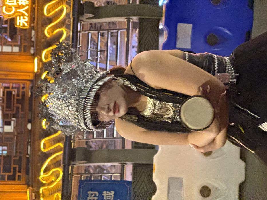A forensic description of the image reveals a nighttime urban scene in Yuzhong District, China, centered on a female individual in traditional ethnic attire. The subject, a woman with dark hair and a neutral expression, faces slightly left while looking forward. She wears an elaborate, ornate silver headdress composed of layered metallic plates and small, hanging reflective rods that form a fringe over her forehead. Large silver earrings and a multi-tiered silver necklace with intricate designs adorn her, complementing her black sleeveless top and black lower garment, which features visible white patterns. Her arms are crossed at her midsection, holding a small, circular hand drum or tambourine with a light-colored skin head and a dark rim.
The background features a building with traditional Chinese architectural elements, brightly illuminated by orange neon tubing outlining decorative curves. Partially visible Chinese characters, including "C位" (center position) and "无人机" (drone), are illuminated in orange neon in the upper right. To the subject's left, a blue sign with white Chinese text is partially visible. White and blue plastic traffic or crowd barriers are present on either side of the subject, suggesting a public or delineated space. The scene is lit by artificial lights, indicative of an evening or nighttime setting. No other individuals are visible, and no direct interactions are depicted beyond the subject holding the drum.